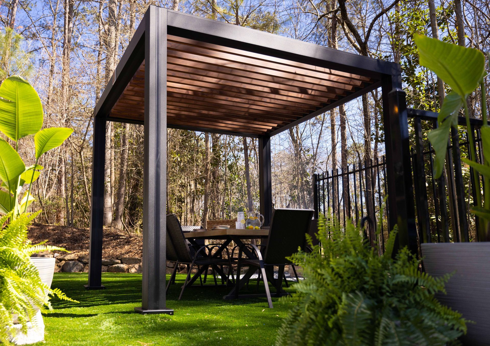 Pergola with dining set on artificial turf in a wooded backyard.