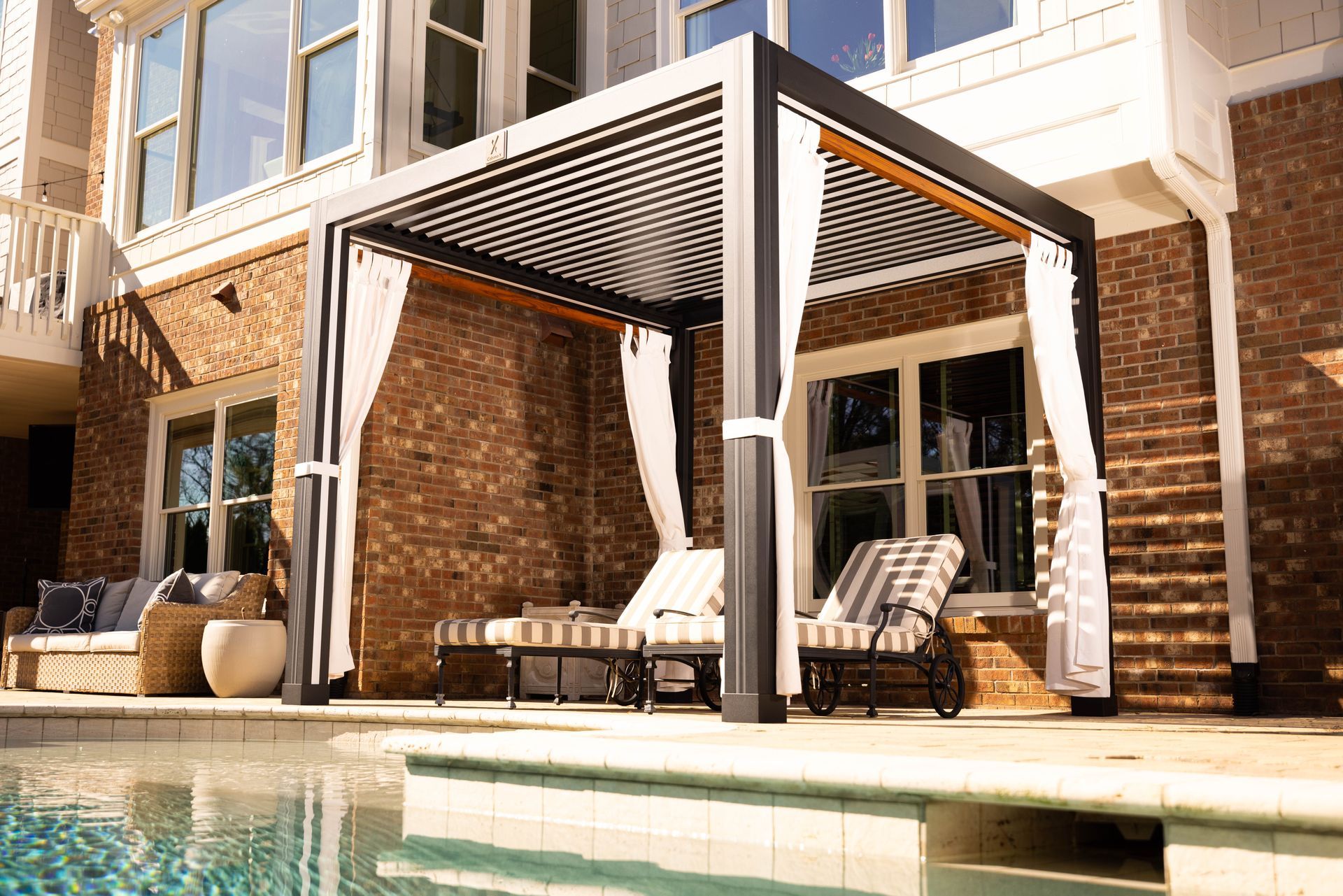 Pergola with curtains next to a pool and brick house with outdoor furniture.