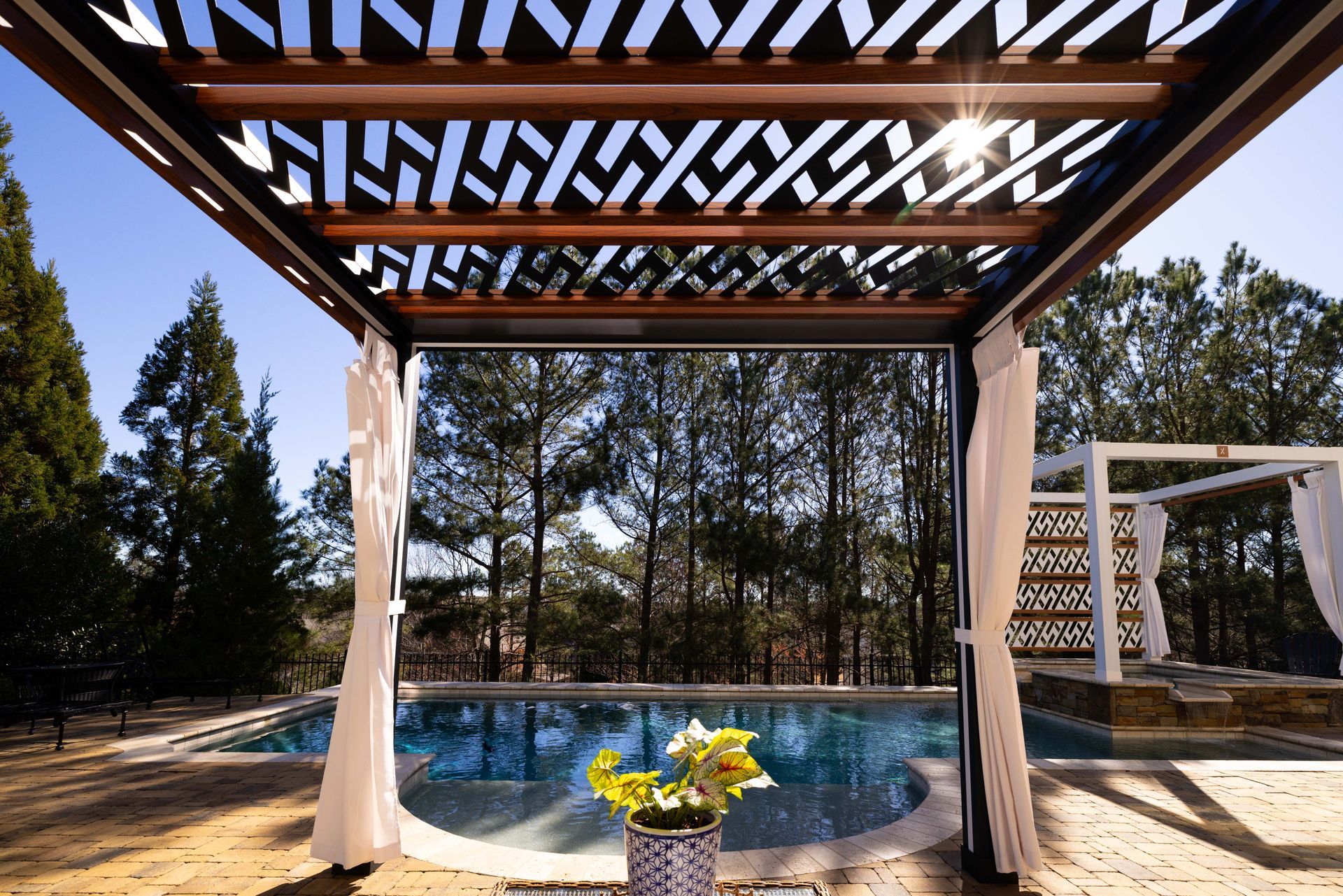 Pergola with intricate overhead design over a pool, white curtains, and trees in the background.