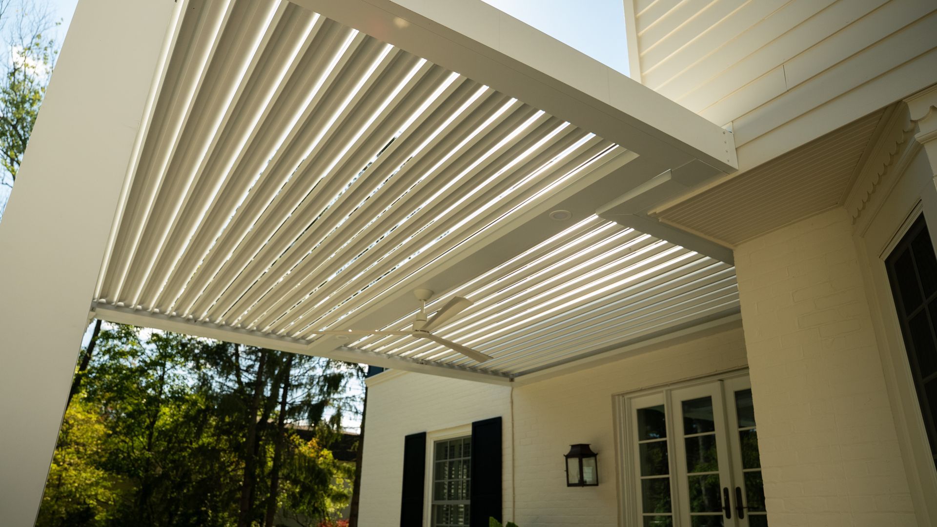 White slatted pergola over a porch with a ceiling fan. Sunny, outdoor setting.