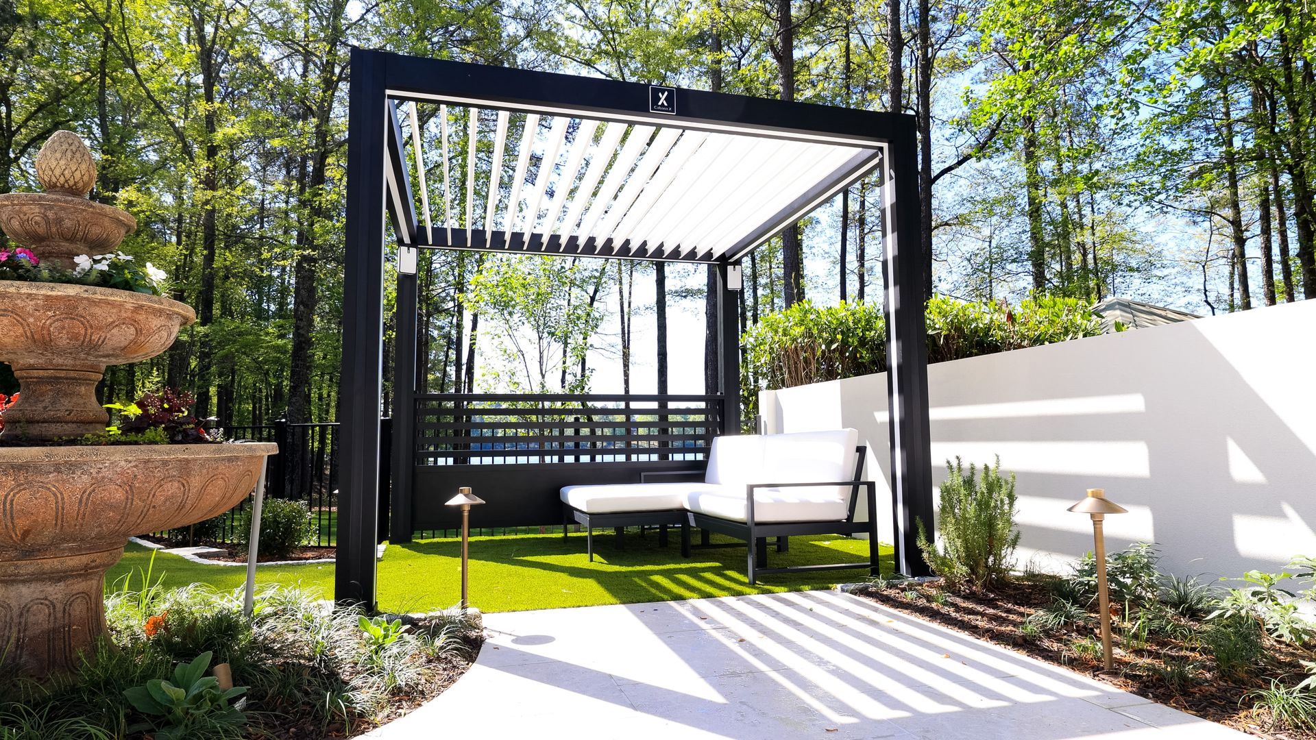 Black pergola with white louvers over outdoor seating on a small green lawn; fountain and white wall backdrop.