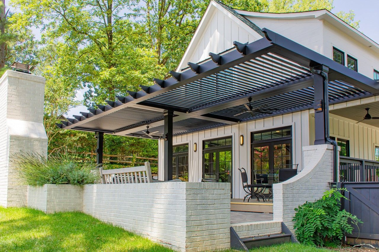 A modern white farmhouse with a black pergola-covered patio, surrounded by greenery.