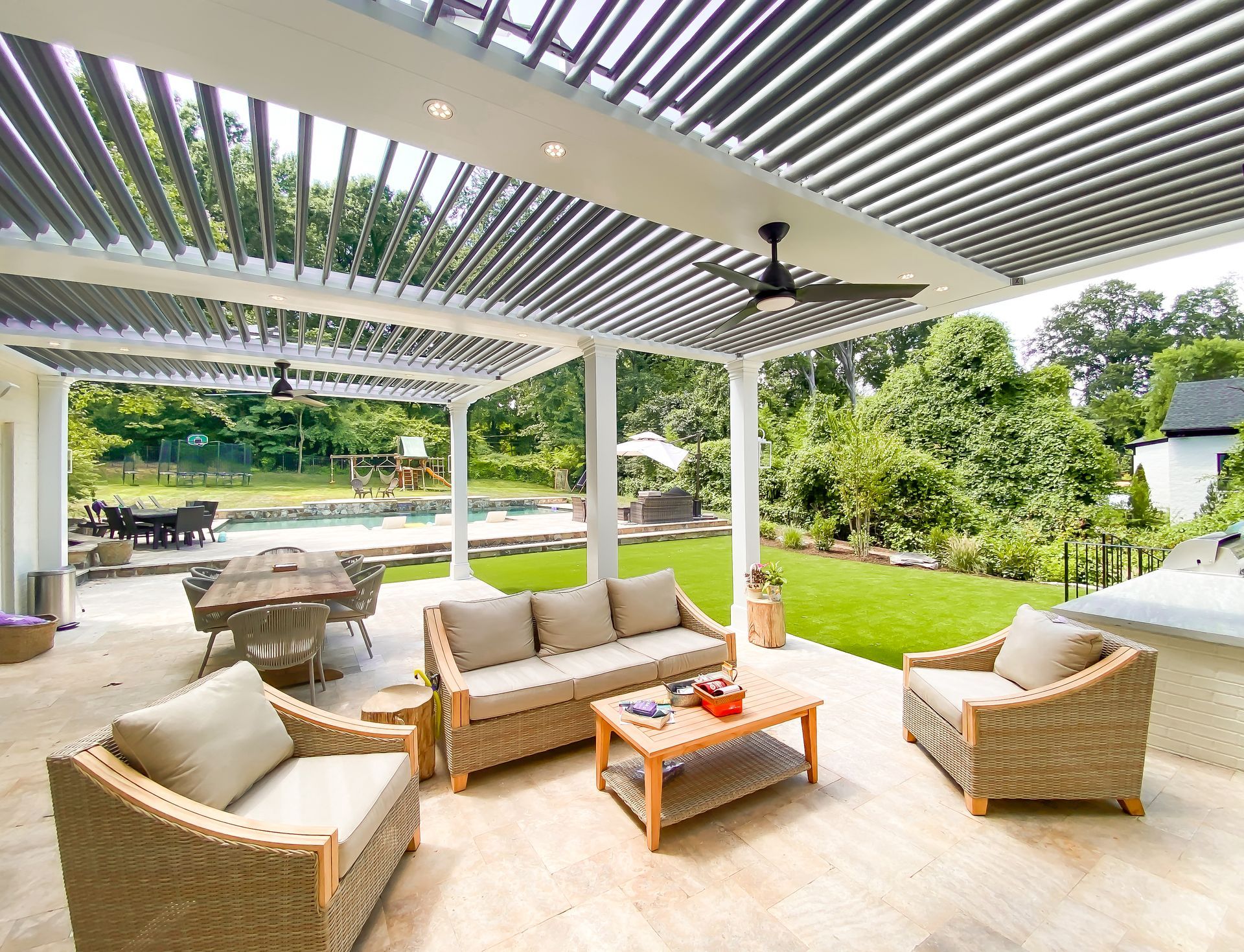 Patio with wicker furniture under a slatted pergola, overlooking a lawn and trees.