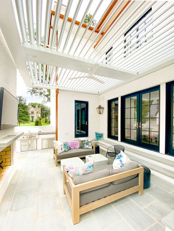 Outdoor patio with light gray couches, white pergola, blue-trimmed windows, and stone floor.