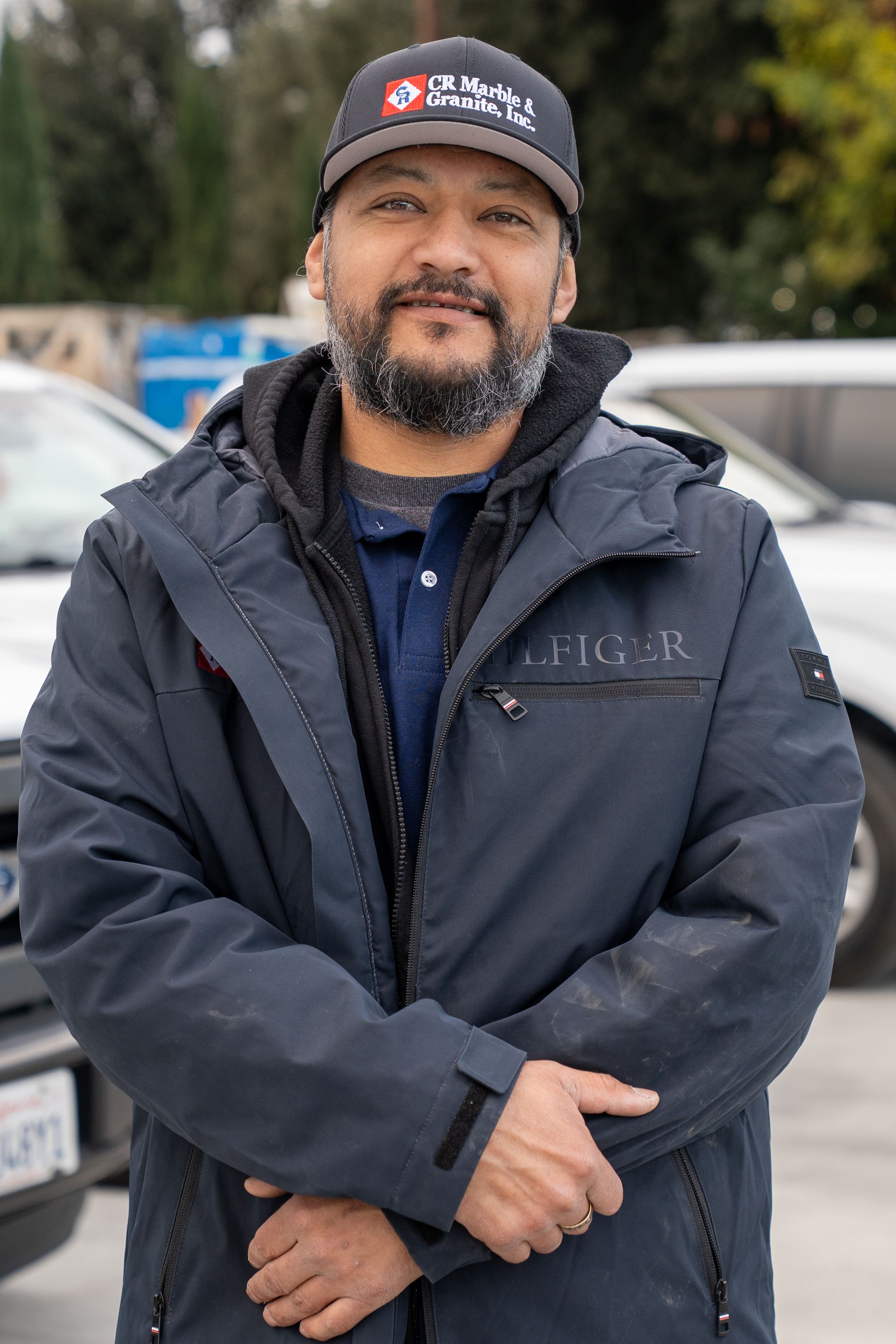A man with a beard wearing a hat and a jacket is standing in front of a car.