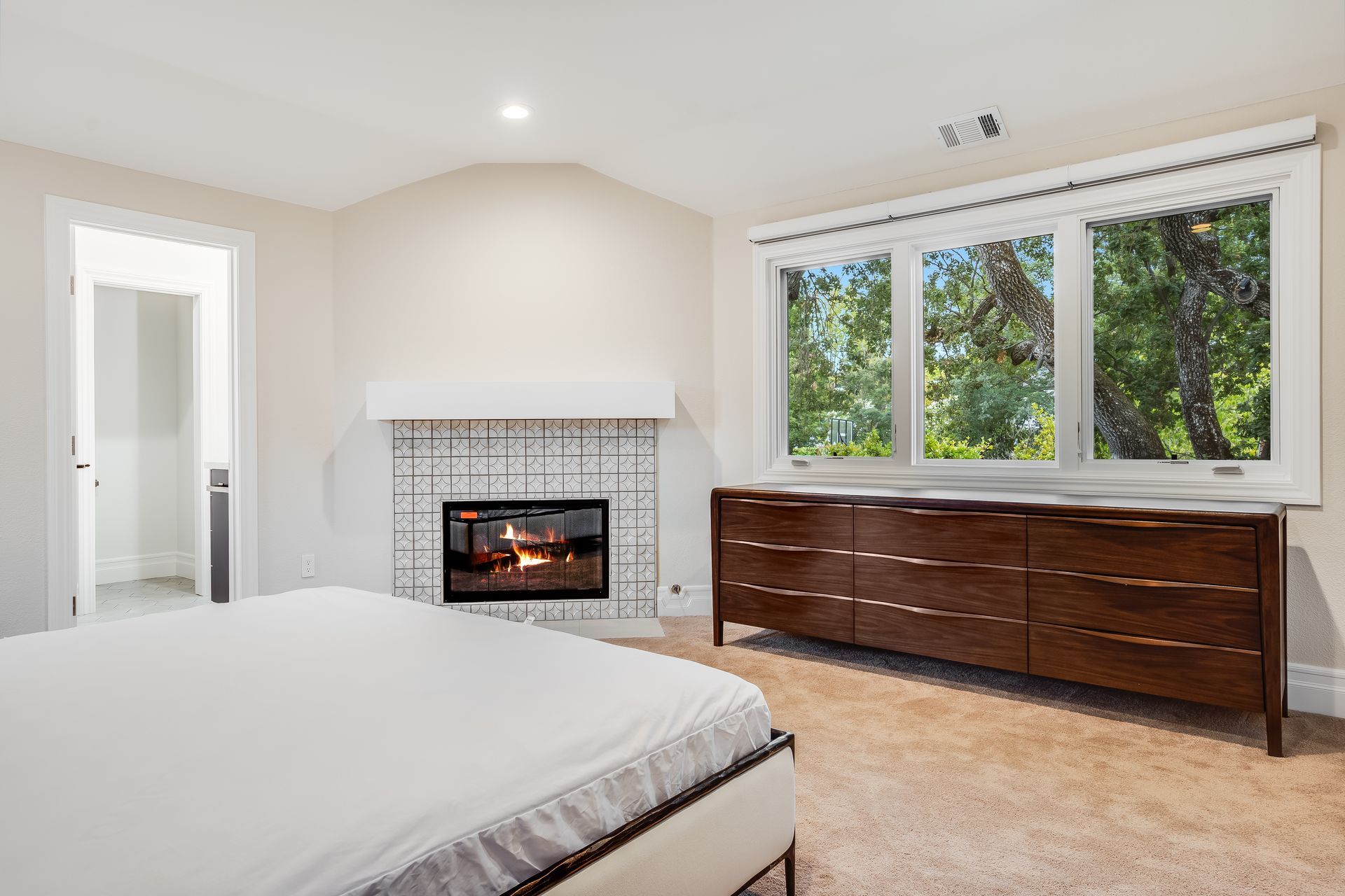 Bedroom with fireplace, large window, wooden dresser, and bed. Fireplace lit. Bright interior.