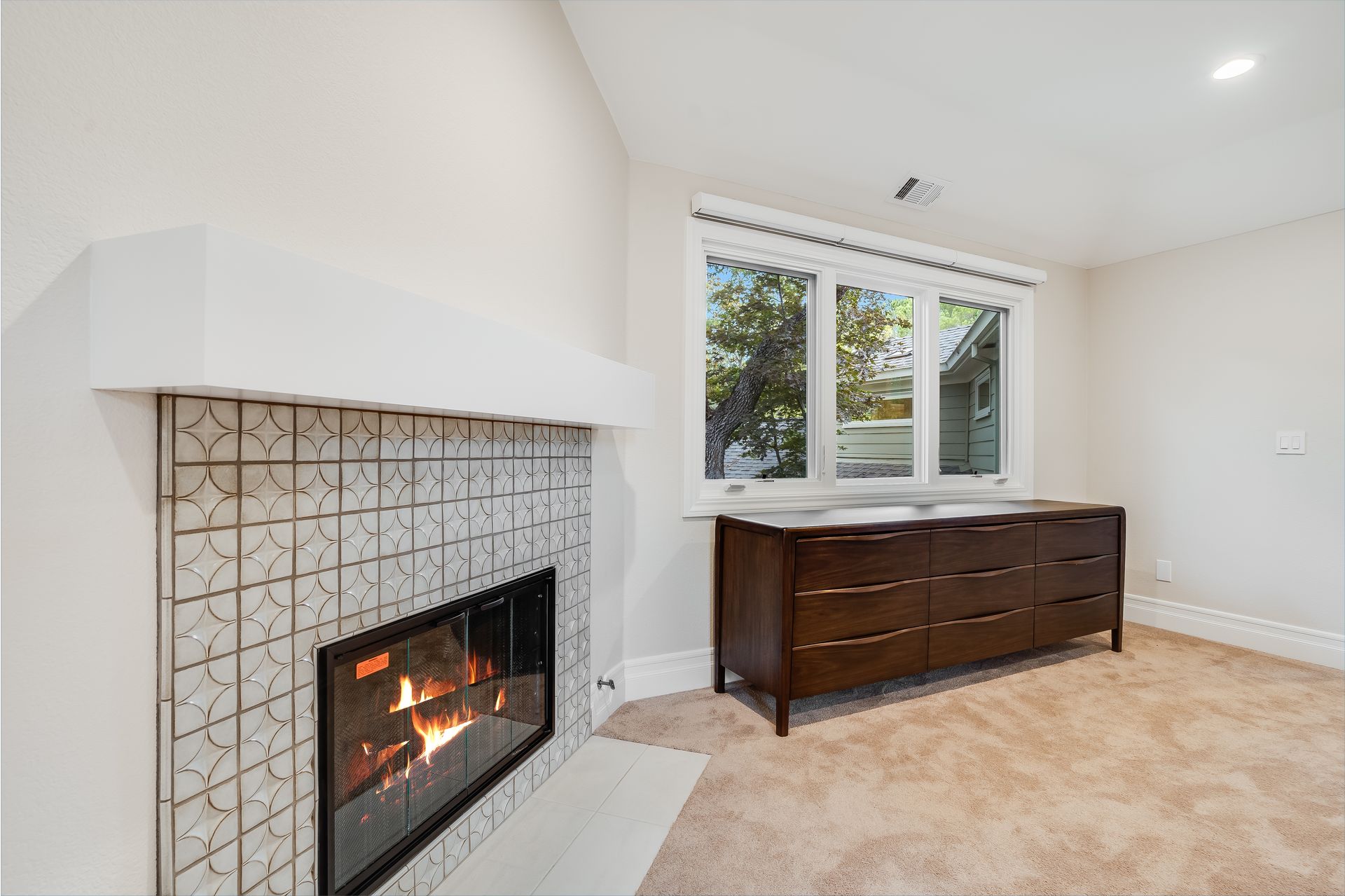 Fireplace with patterned tiles, brown dresser by a window in a room with neutral walls and carpet.