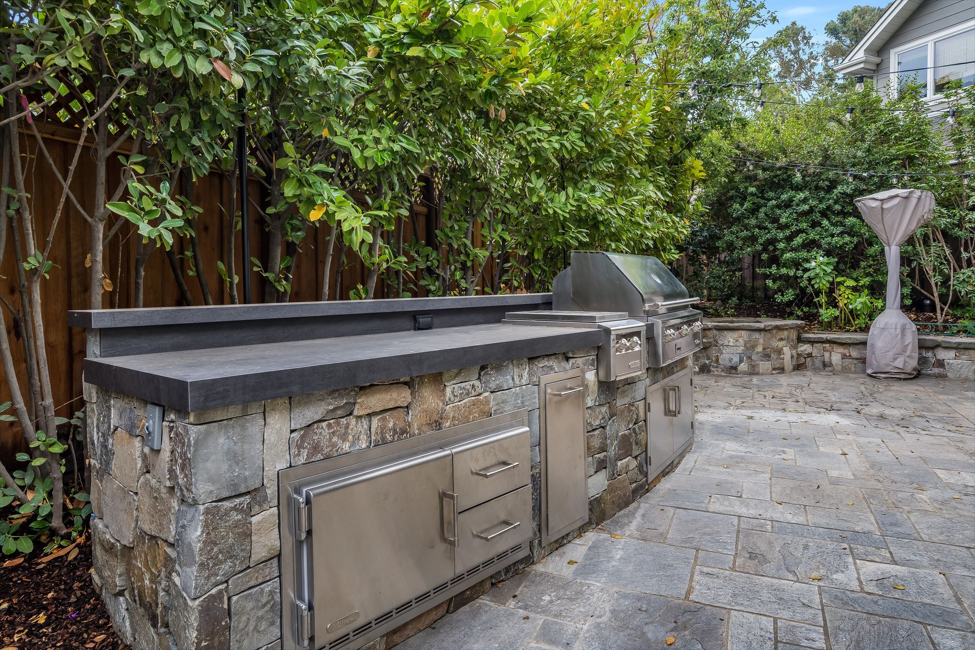Stone-covered outdoor kitchen with stainless steel grill, cabinetry, and a patio, surrounded by greenery.