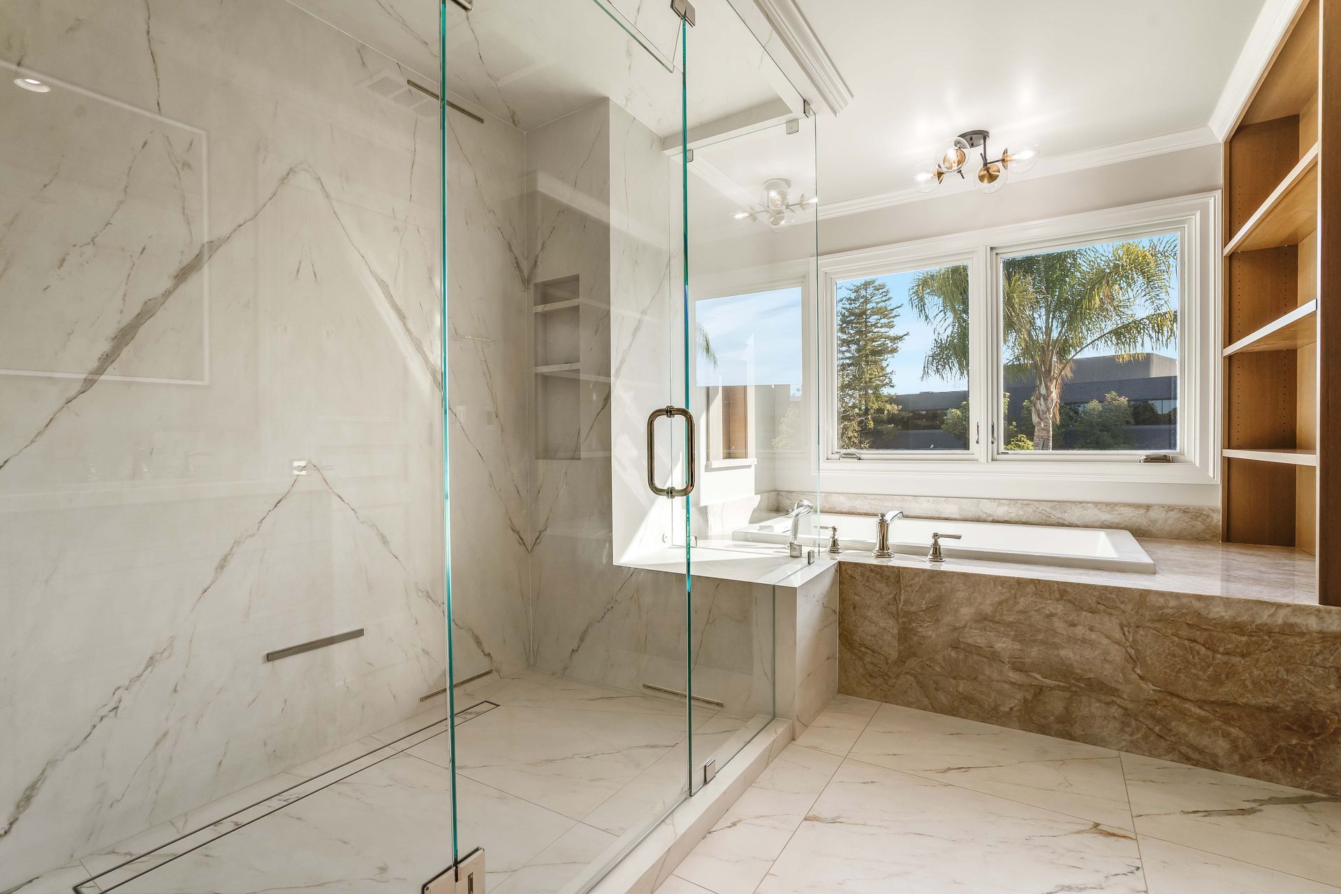 Luxurious bathroom with marble walls and glass shower. Bathtub, window, and built-in shelving are also present.