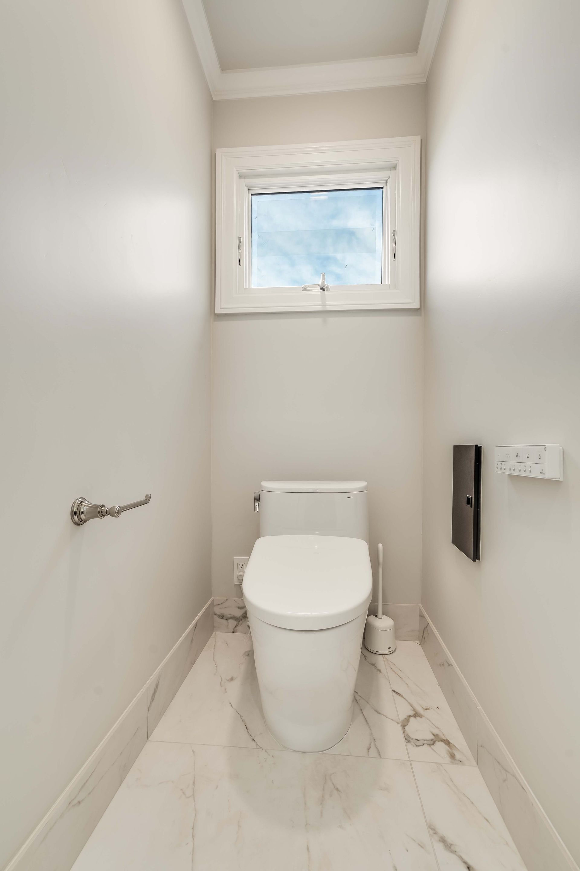 Small, white bathroom with a toilet, window, marble floor, and paper towel holder.