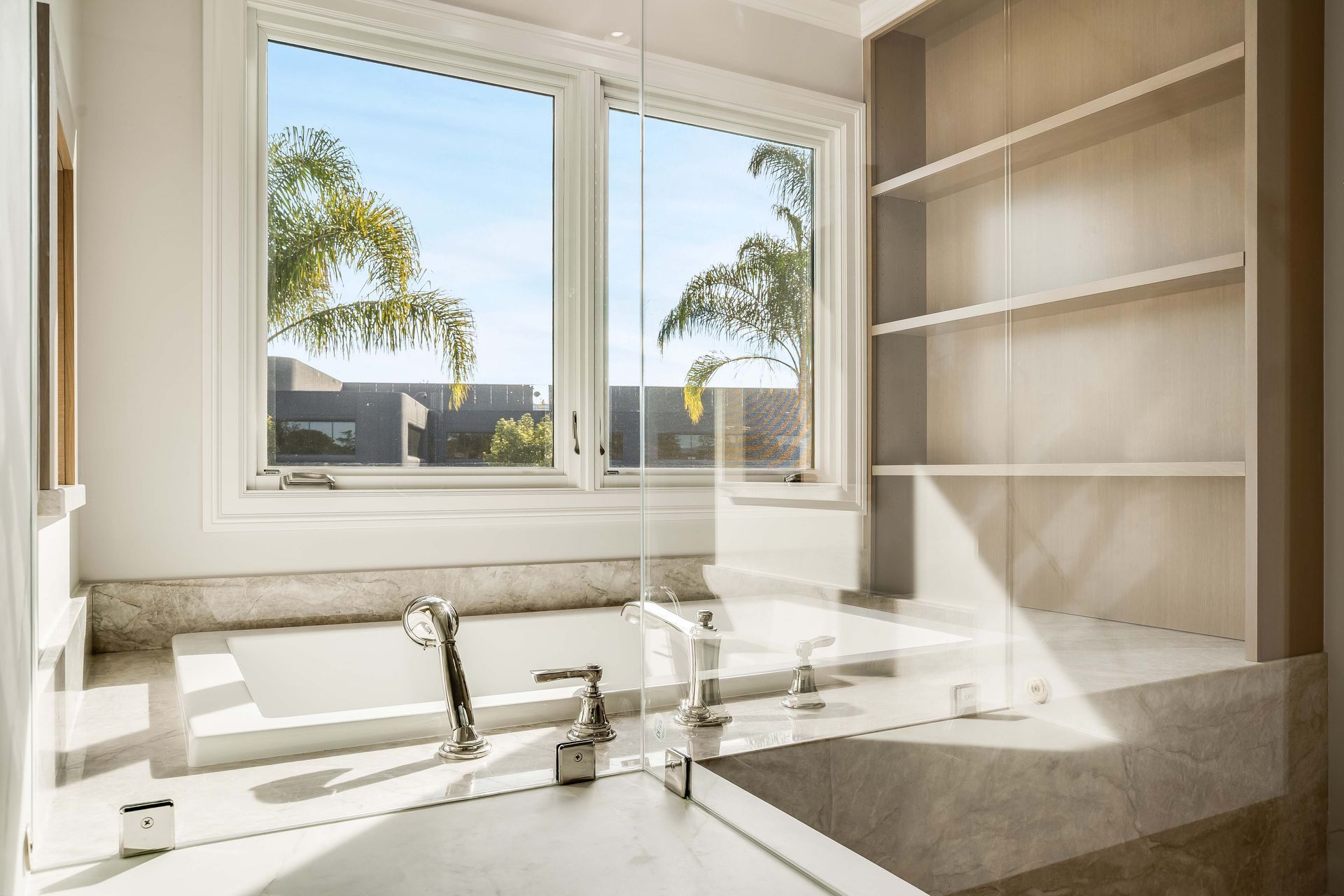 Bathroom with a marble counter, soaking tub, window with view of palm trees, and built-in shelves.