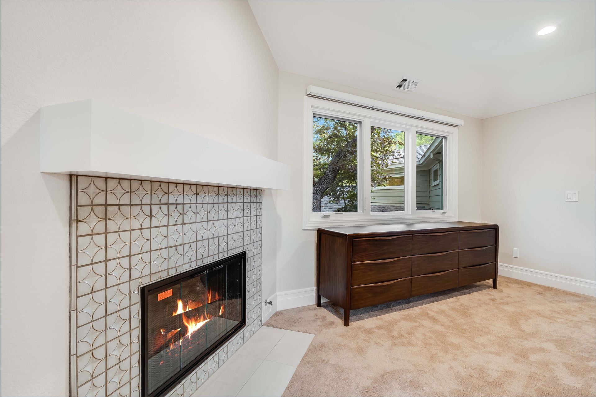 Fireplace with patterned tile, lit fire, and a dark wood dresser in a room with a large window.