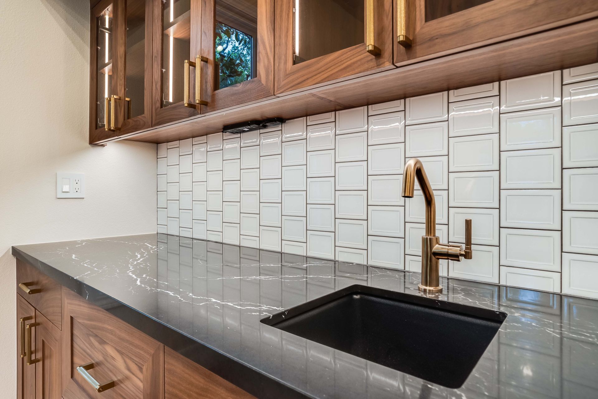 A dark kitchen sink with gold faucet, black countertop, and white tile backsplash. Wooden cabinets with gold handles.
