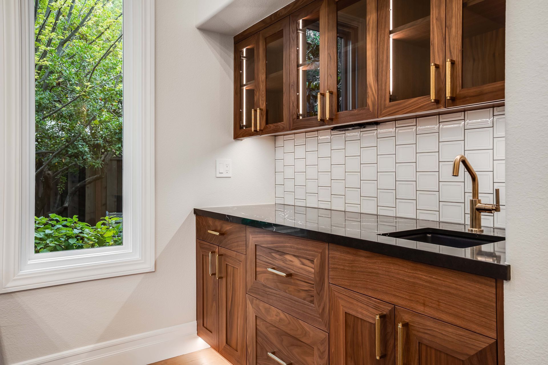 A small home bar with dark wood cabinets, black countertop, white tile backsplash, and a window overlooking a green yard.