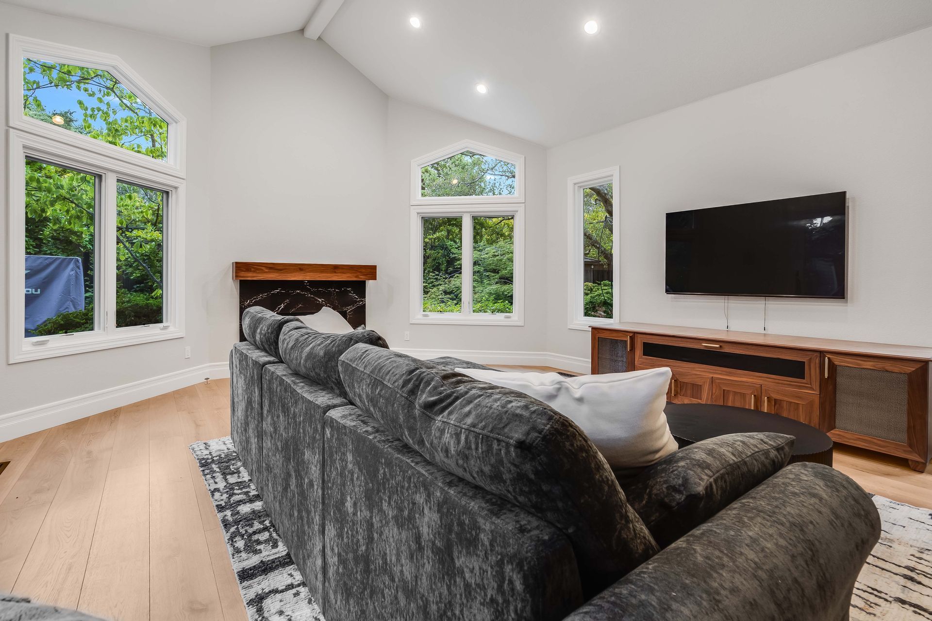 Living room with a large gray sectional, TV, and large windows with a view of greenery.