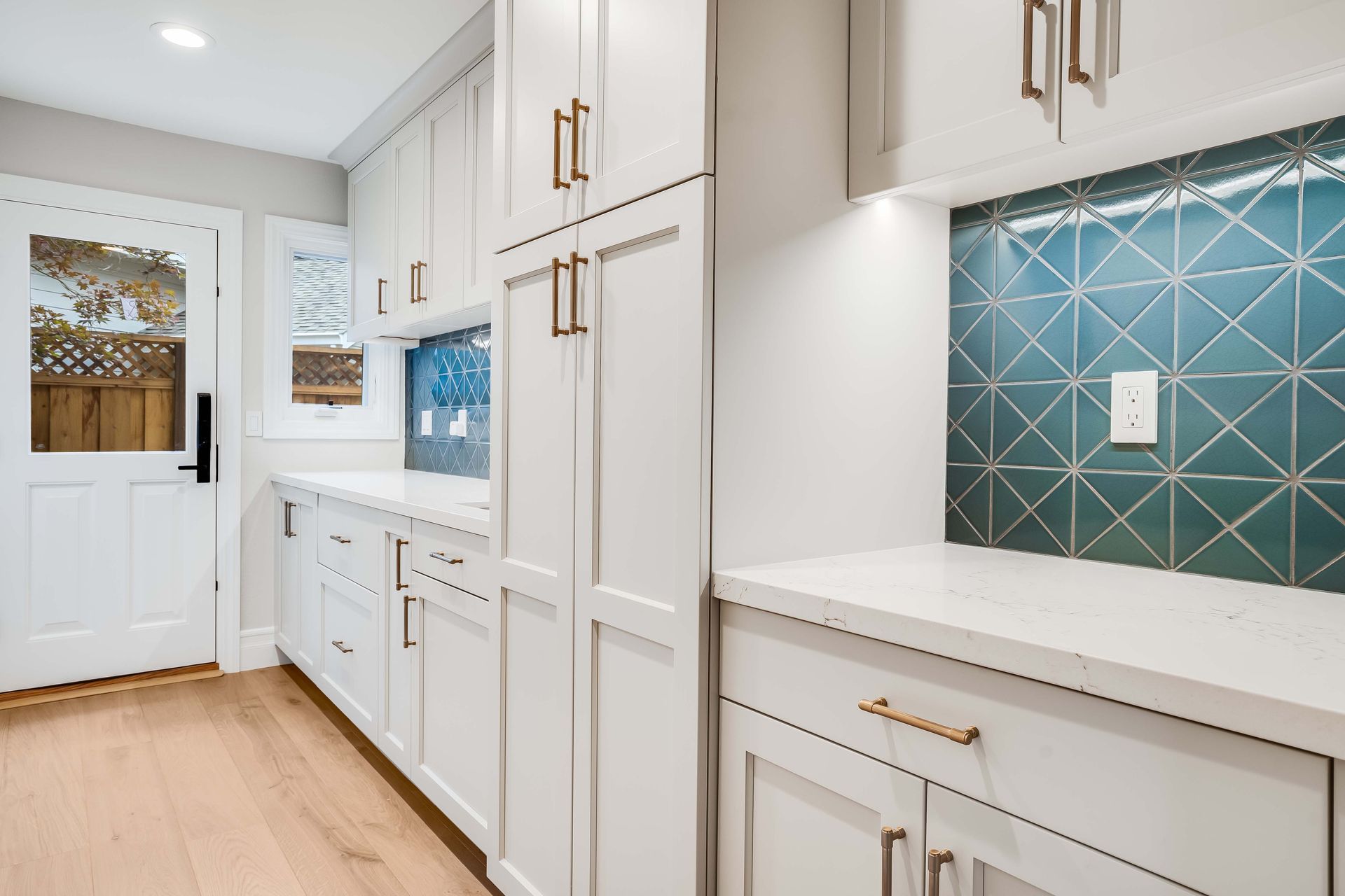 White kitchen cabinets with gold hardware, teal tile backsplash, and light wood flooring.