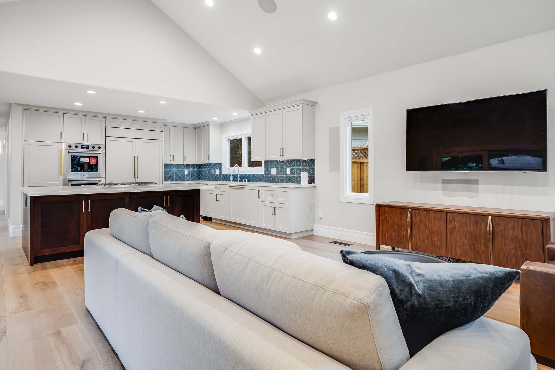 Modern living room with open kitchen, white walls, wood floors, dark wood island, and large TV.