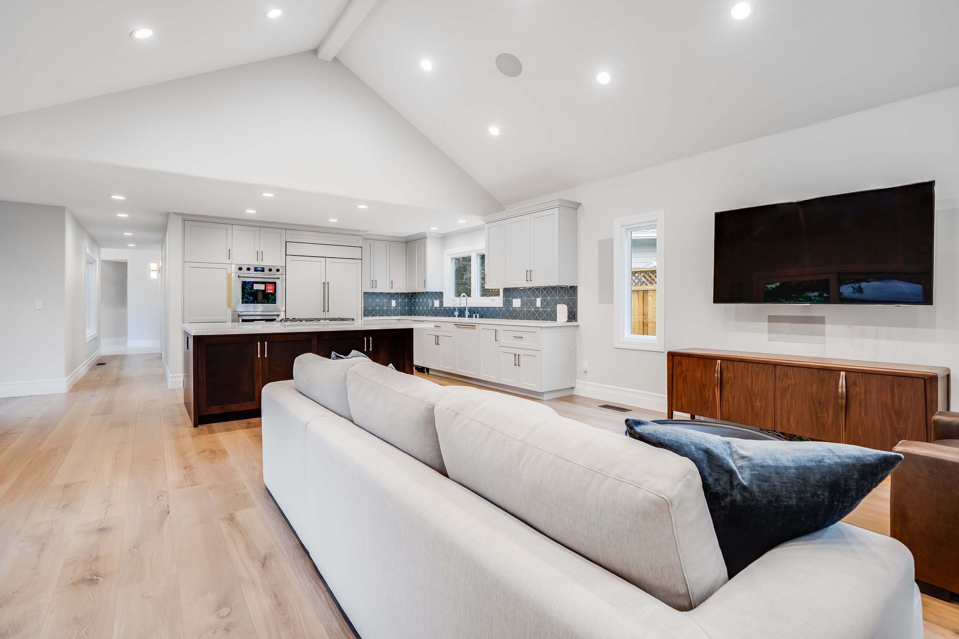 Open-concept living space with kitchen and living room. Neutral colors, wood floors, white cabinetry, dark island, large TV.