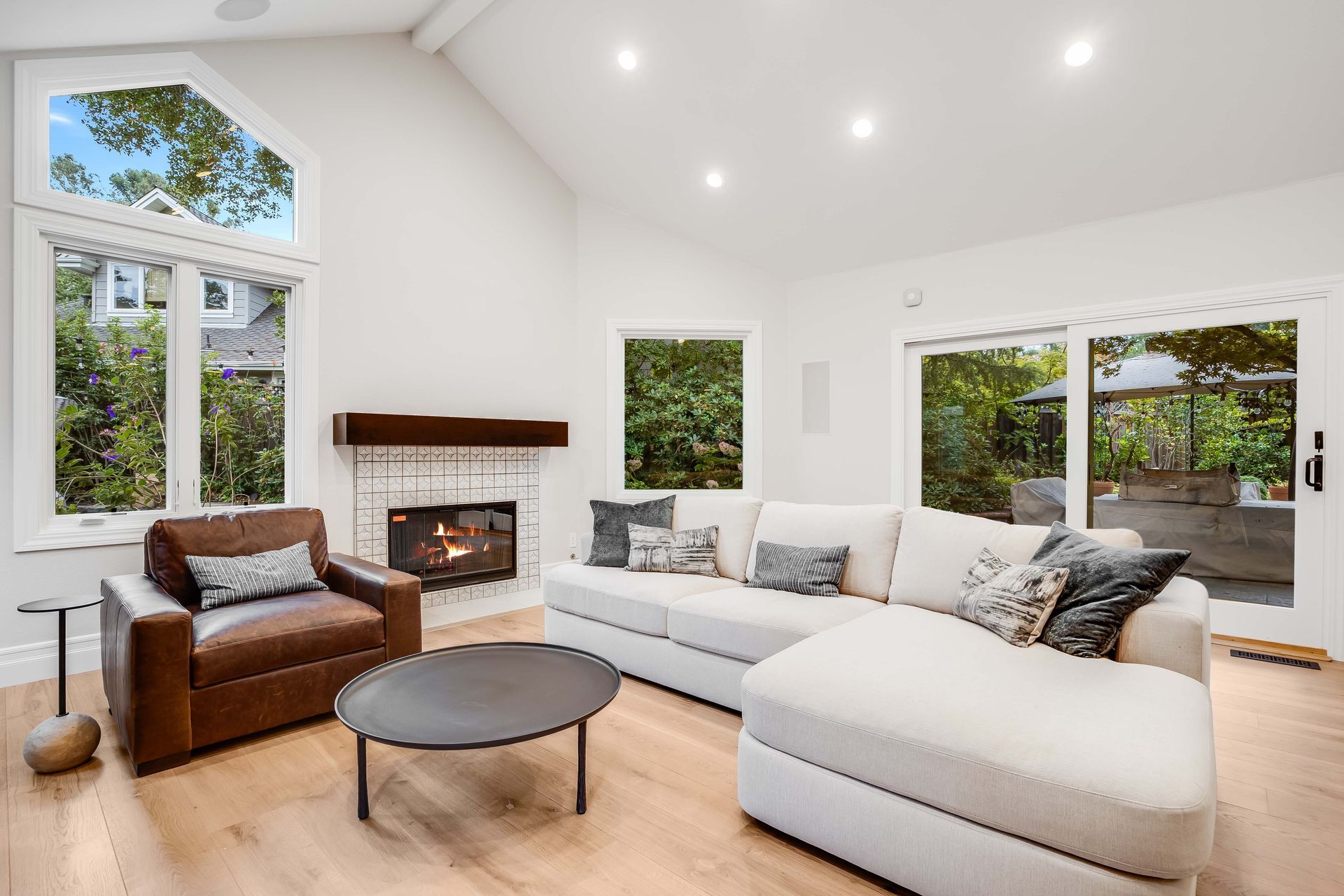 Bright living room with white walls, fireplace, and large windows. Brown leather chair, white sofa, and wood floor.