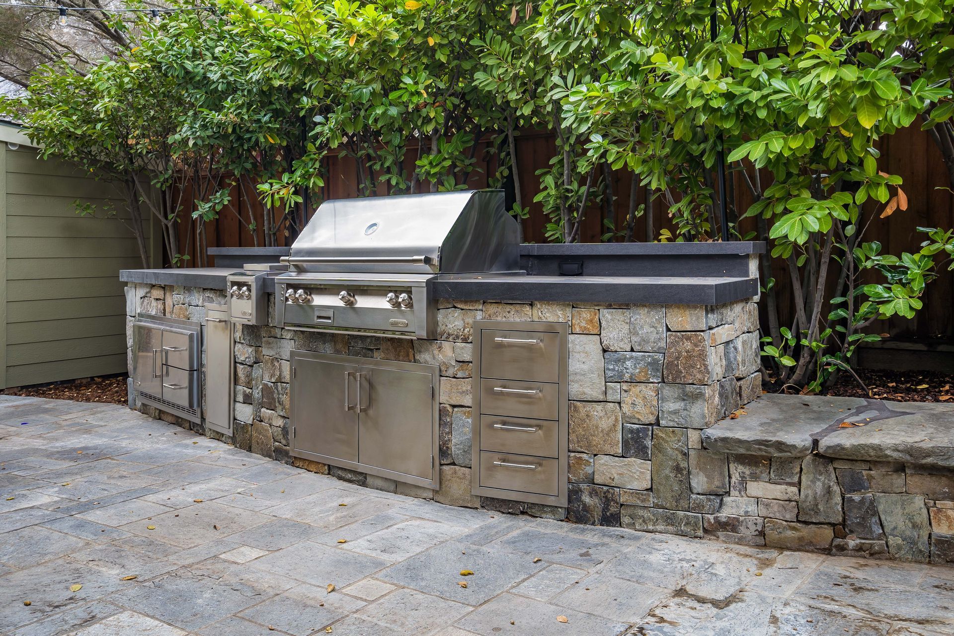Outdoor kitchen with stone facade, stainless steel grill and cabinets, set on a paved patio.