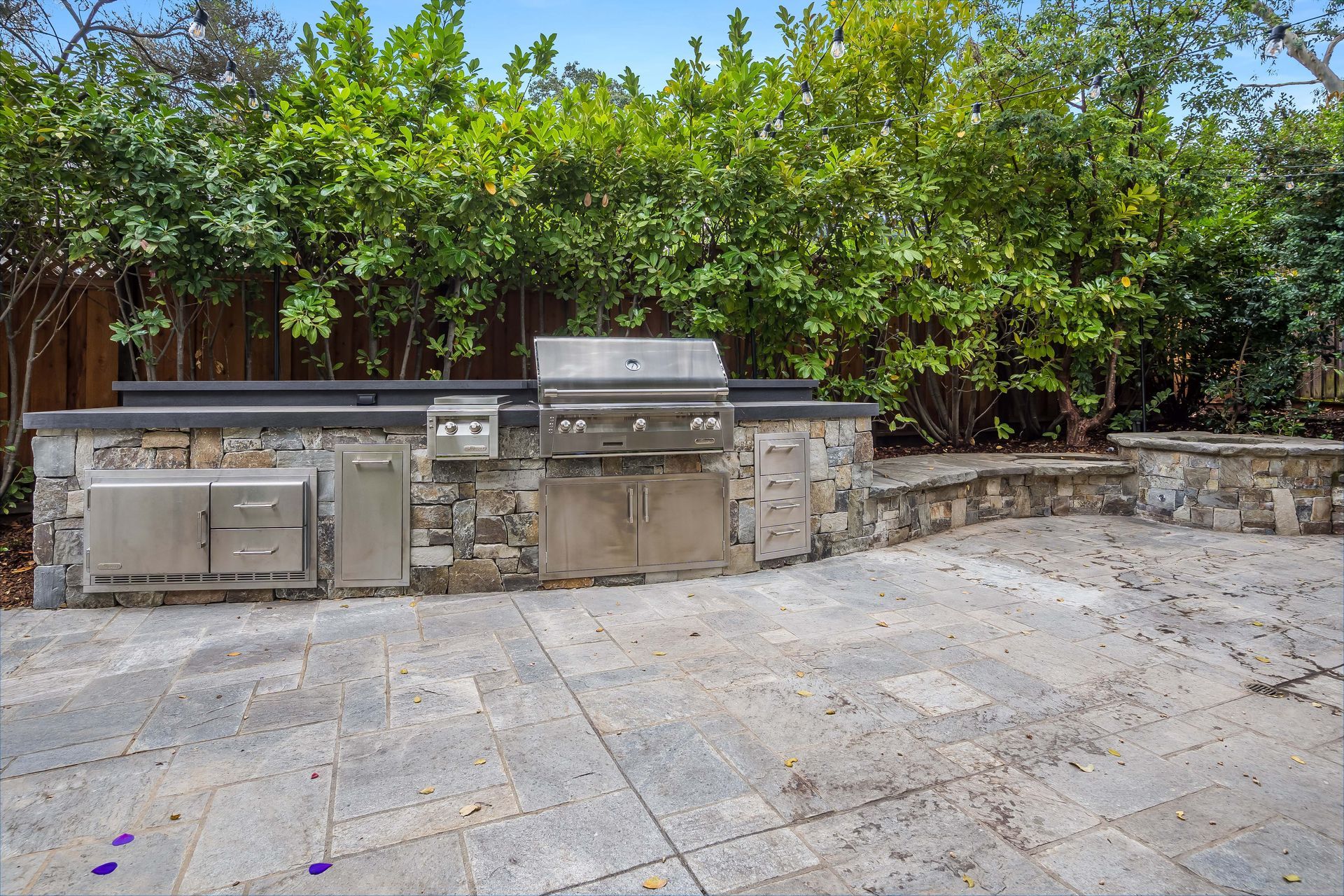 Outdoor kitchen with grill, stone counter, stainless steel appliances, and a paved patio in the background.