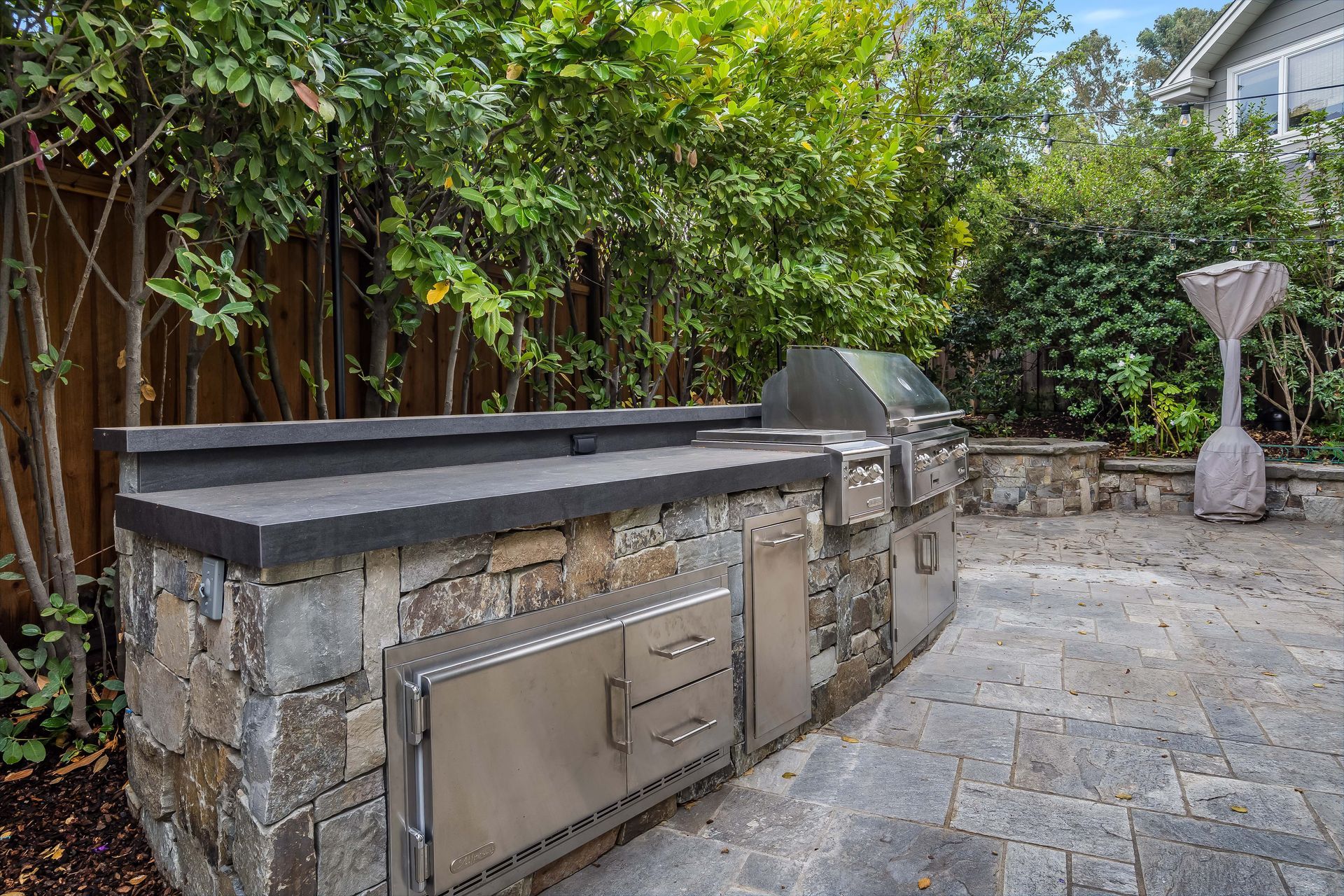 Outdoor kitchen with stone base, stainless steel appliances, and dark countertop on a paved patio.