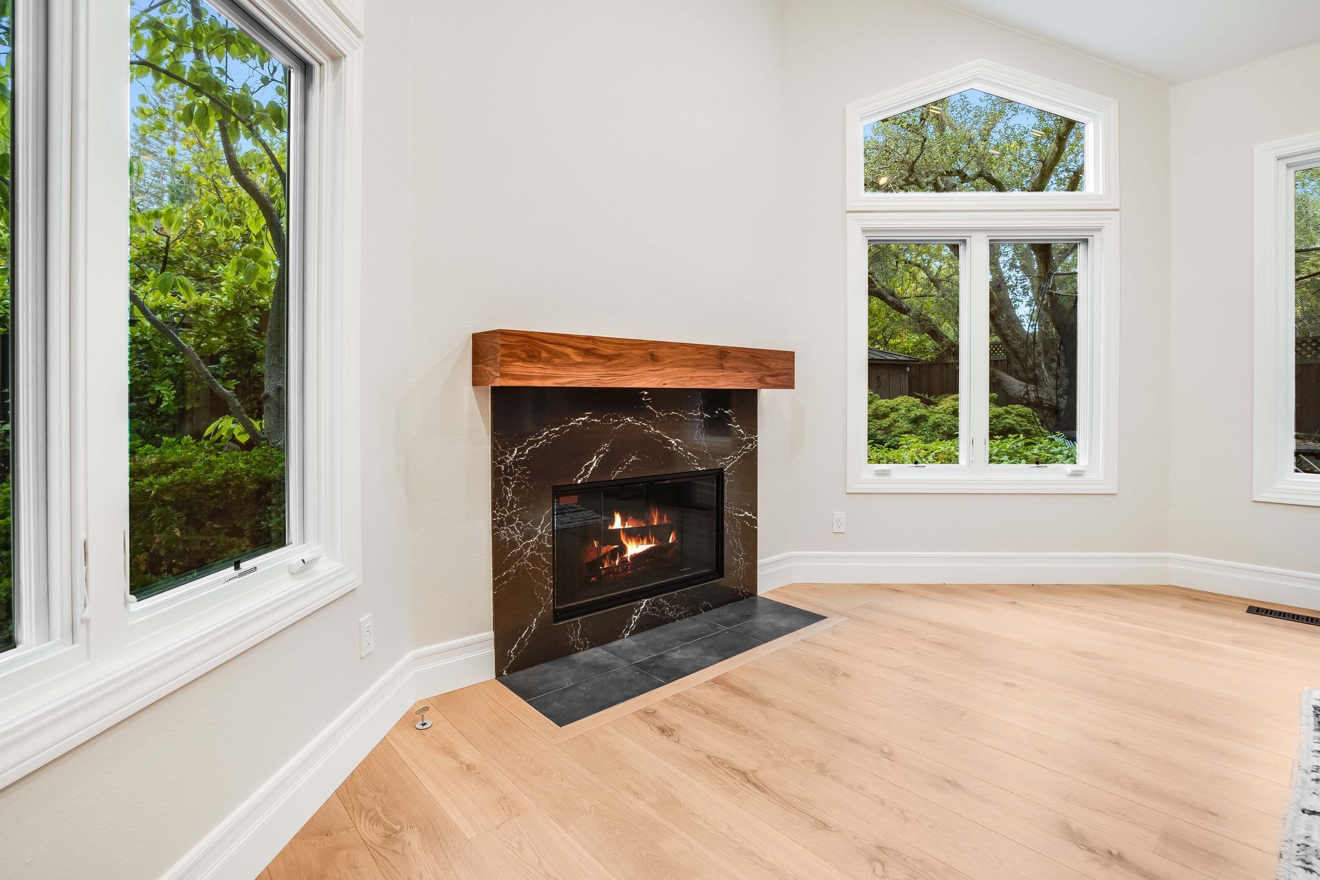 Fireplace with a black marble surround, wood mantel, and windows in a bright room.