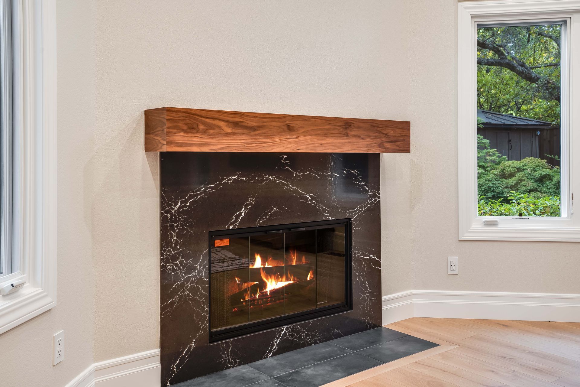 Fireplace with black marble surround, wooden mantle, and flames, next to a window.