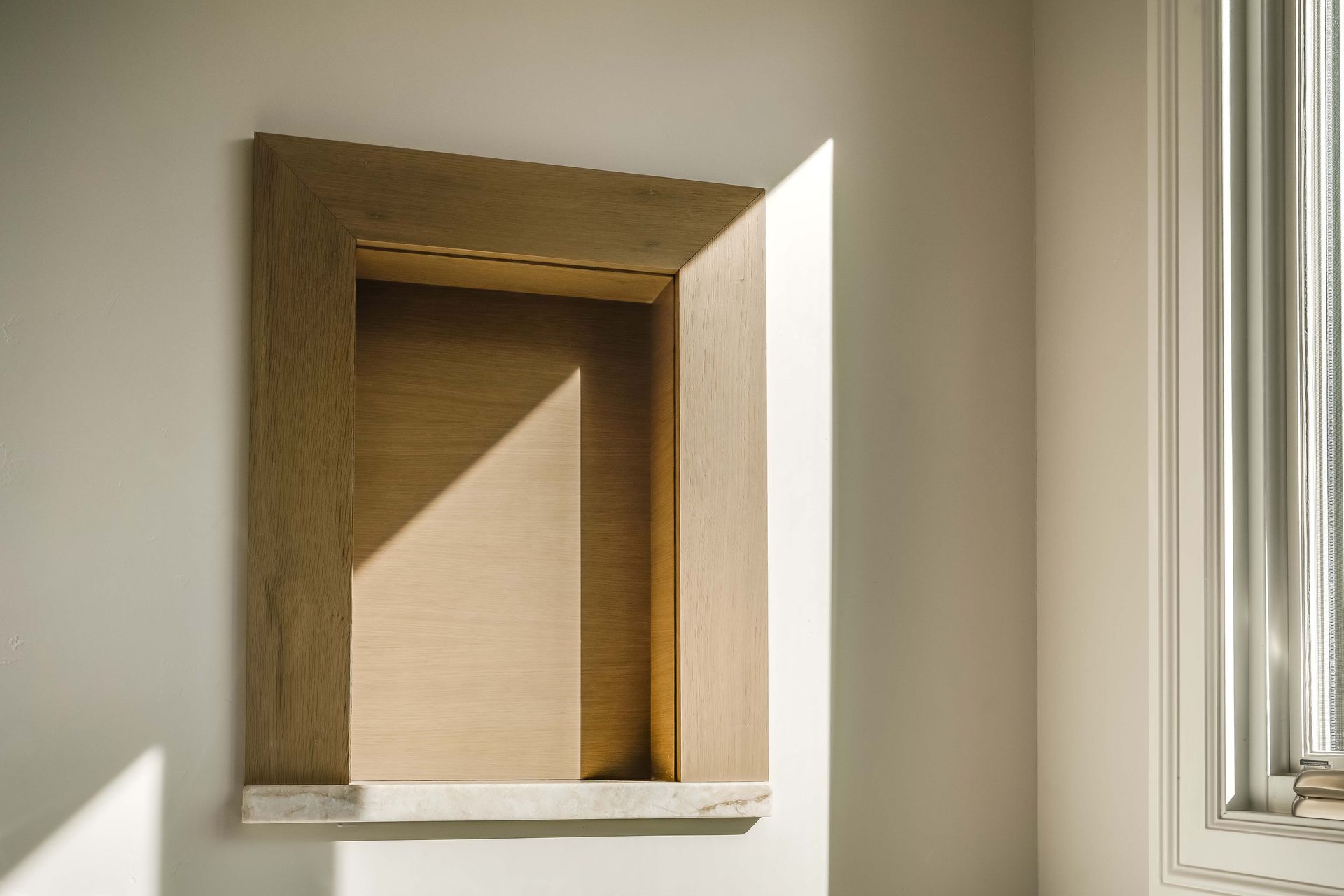 Empty recessed wooden niche on a white wall with sunlight streaming in; a window is to the right.