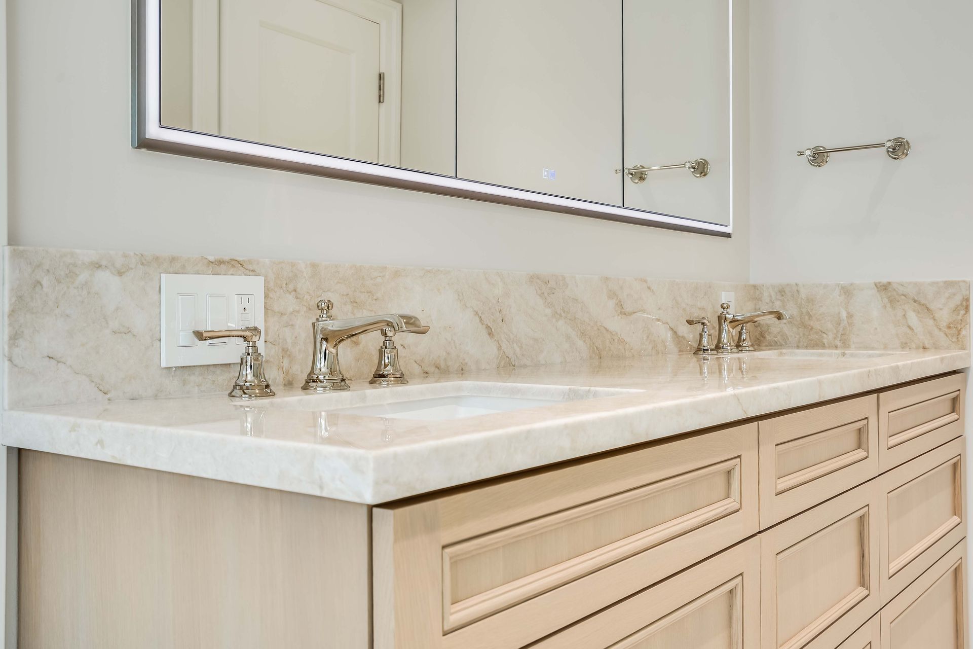 Bathroom vanity with light marble countertop, two sinks, and silver fixtures. Light wood cabinets.