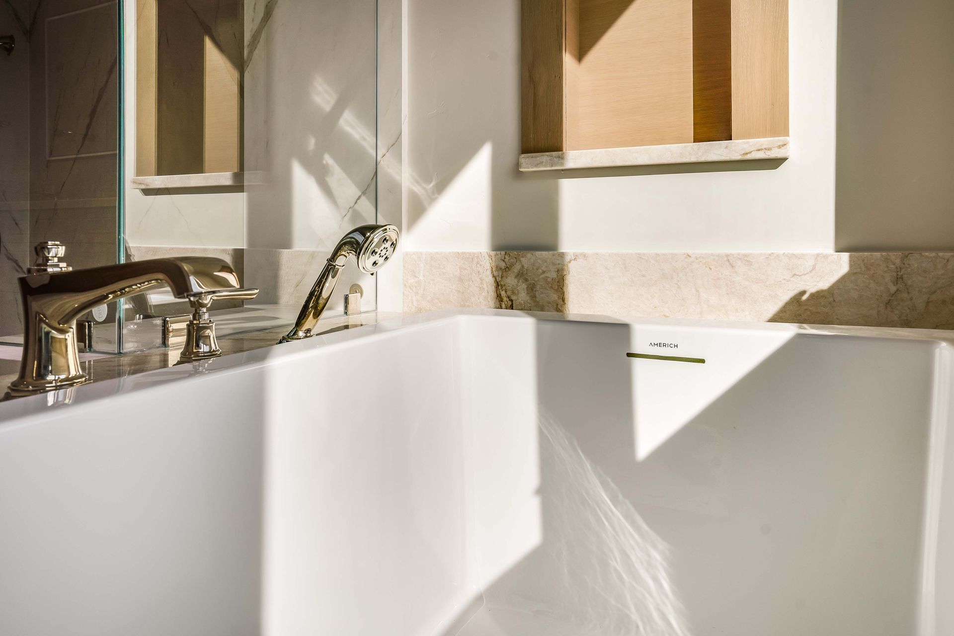Close-up of a white bathtub with gold faucet. Sunlight casts shadows on the tub and wall.