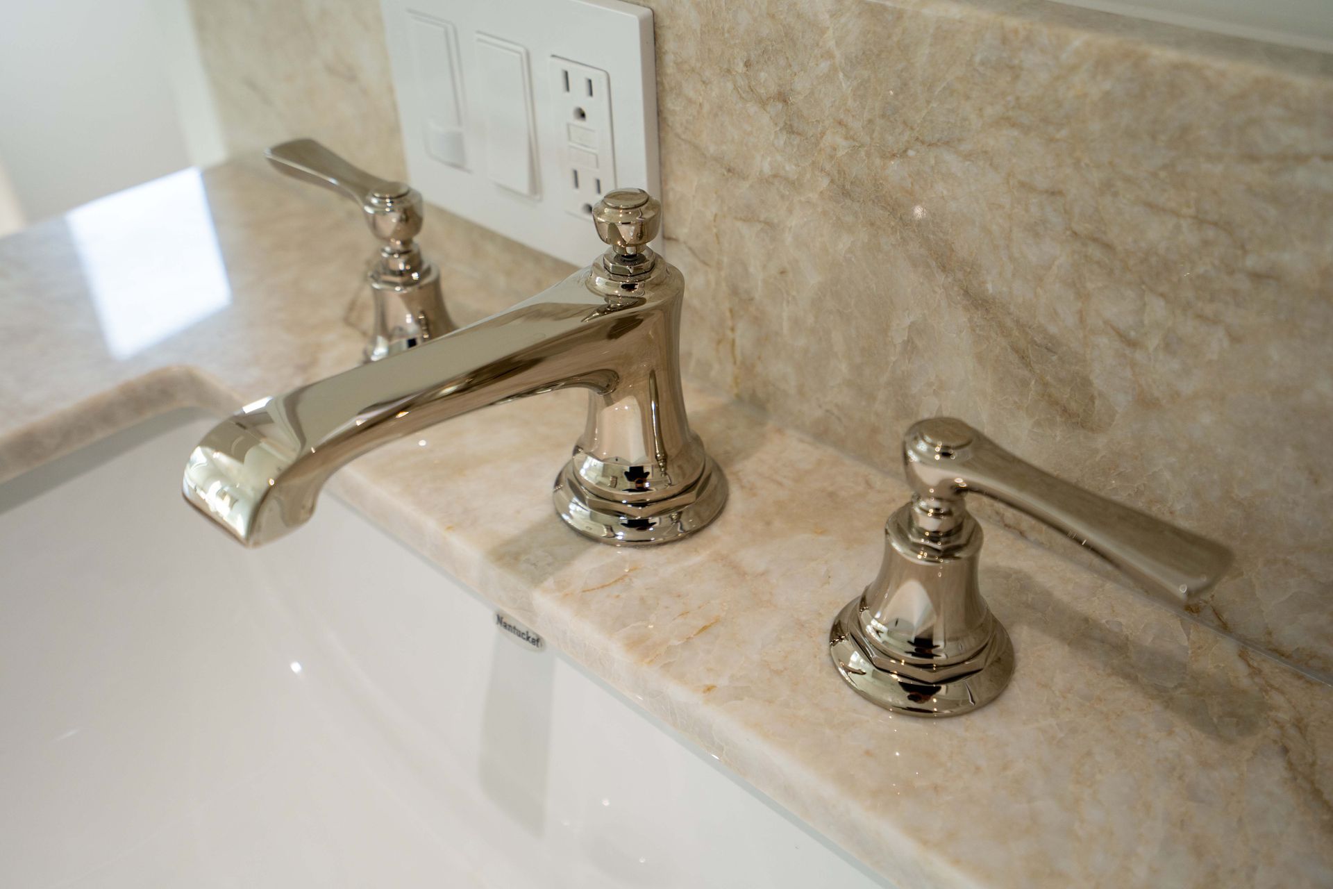 Shiny silver faucet with two handles on a light tan marble countertop. White sink visible on left.