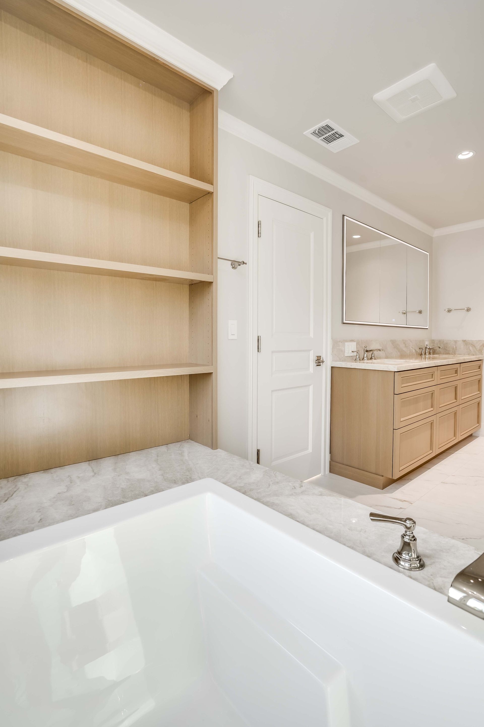 Bathroom with built-in shelves, white bathtub, vanity, and closed door. Light wood and white accents.