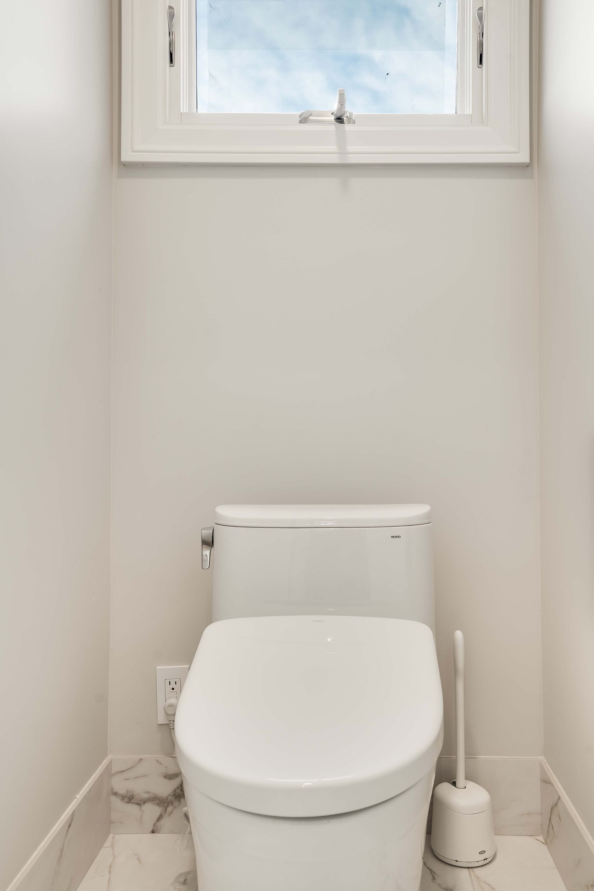 White toilet in a small bathroom with a window and a toilet brush.