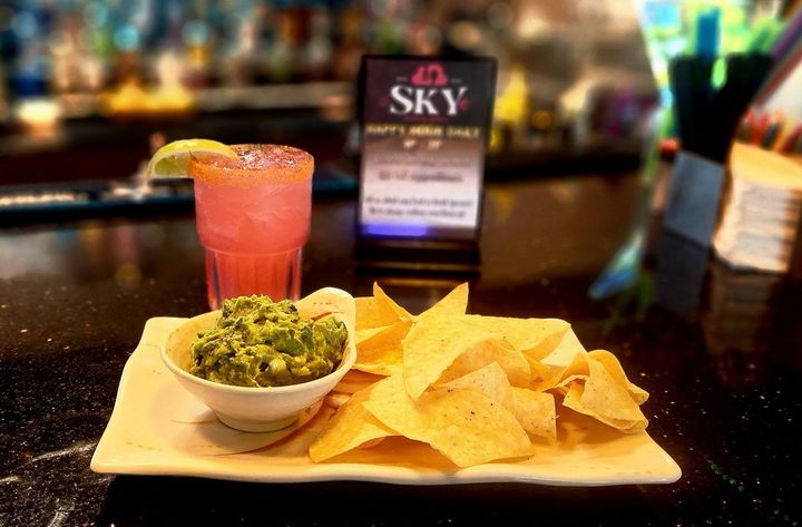 Red cocktail and tortilla chips with guacamole on a tray at a bar, with a blurred drink menu in back