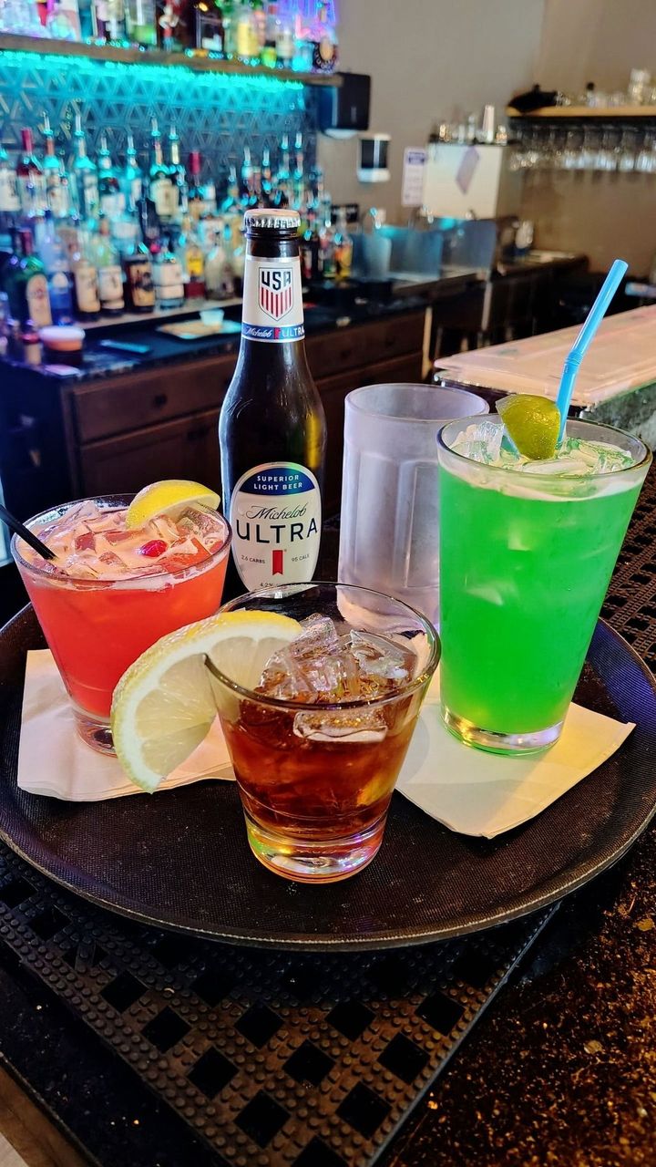 Colorful cocktails and a beer bottle on a bar table, with a neon-lit bar in the background