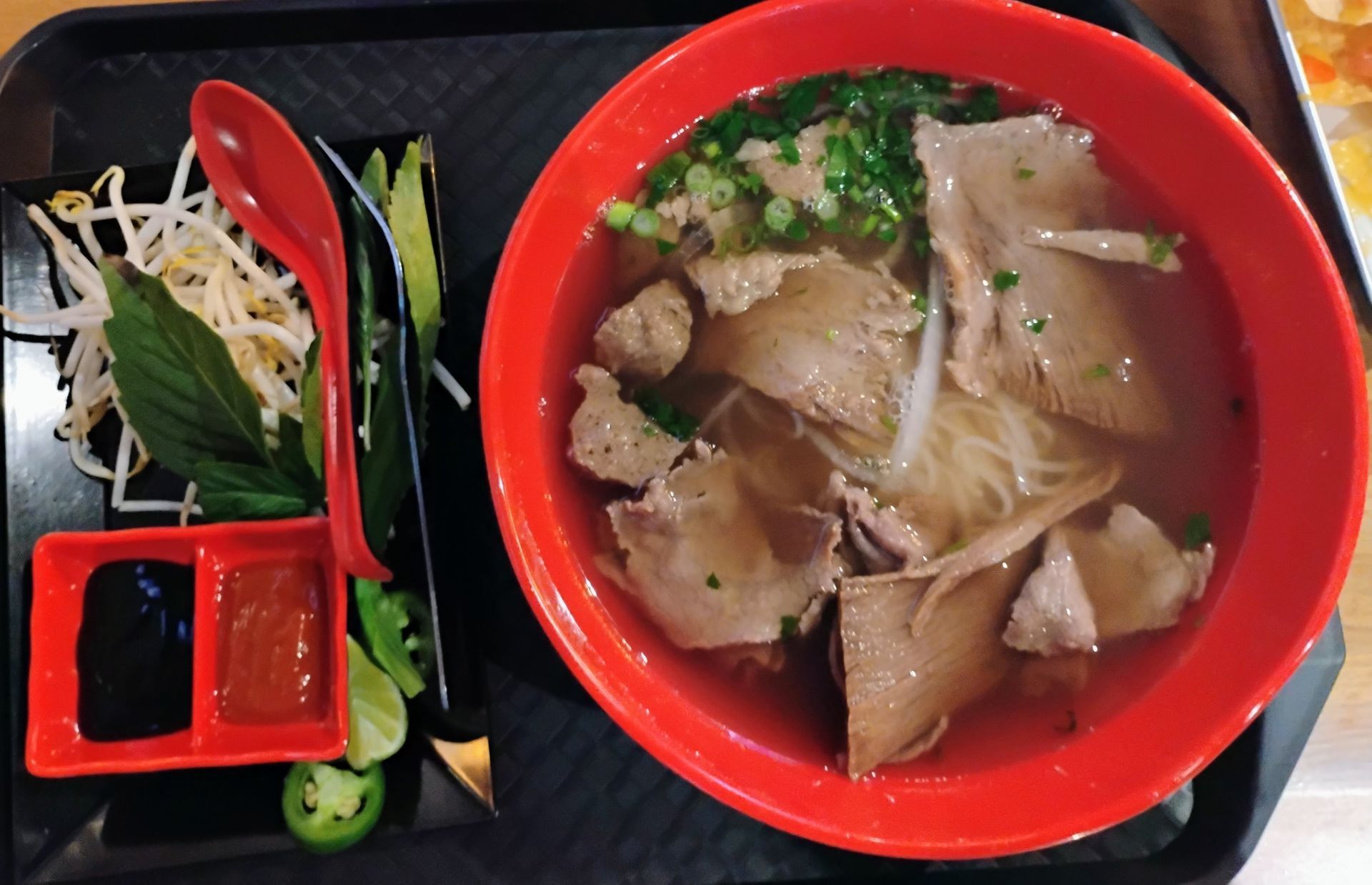 Red bowl of noodle soup with sliced meat, plus small side dishes on a black tray with red utensils.