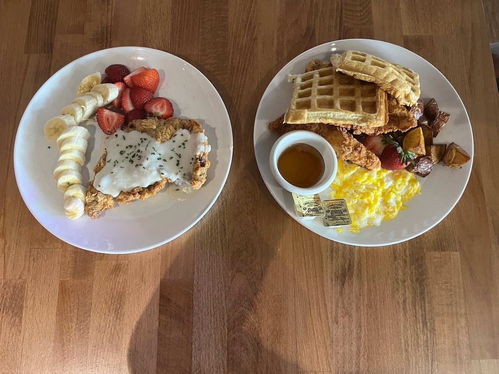 Two breakfast plates on a wooden table with waffles, eggs, fruit, and coffee