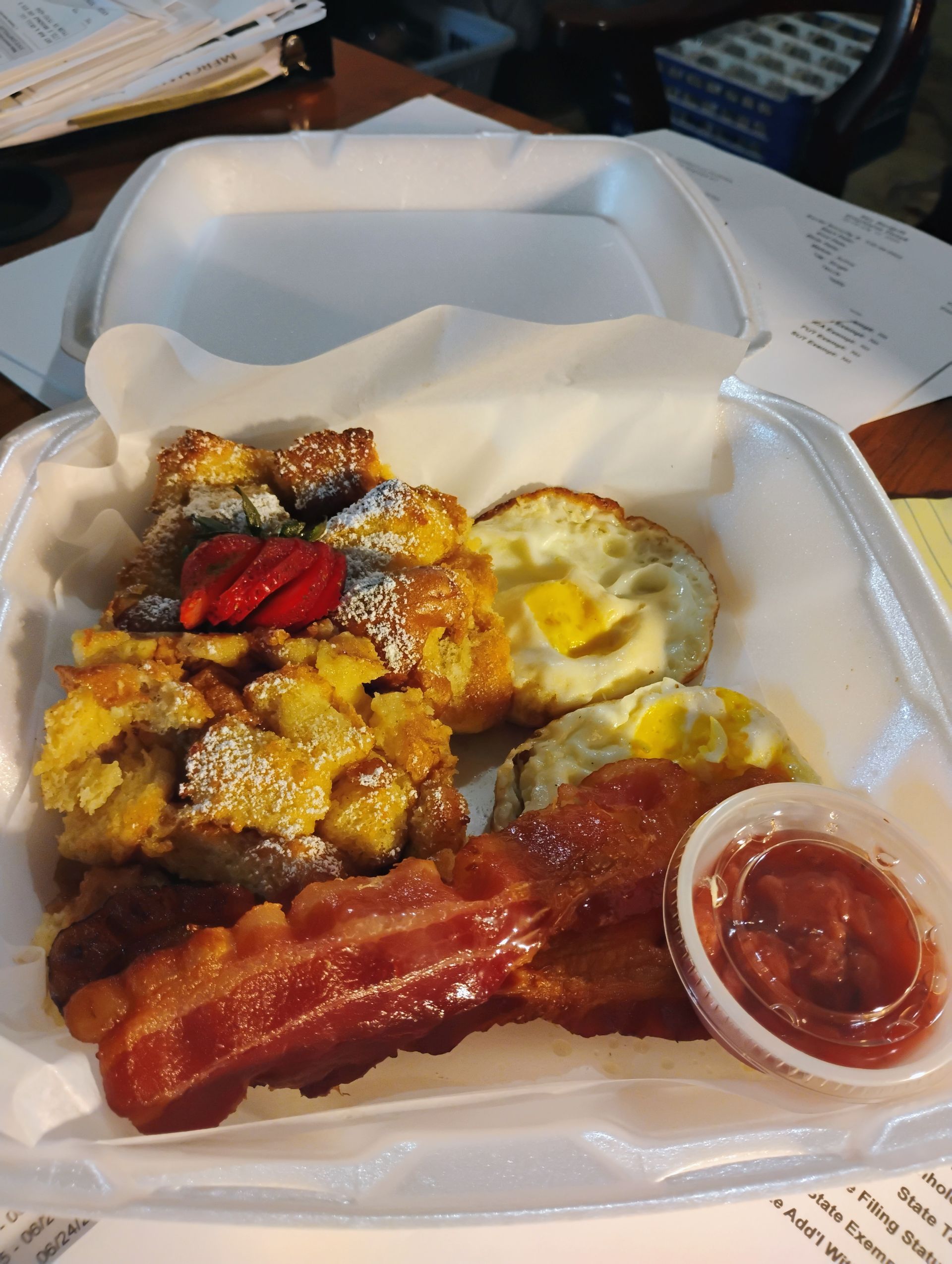 Breakfast in a foam takeout box with pancakes, eggs, bacon, berries, and syrup on a table
