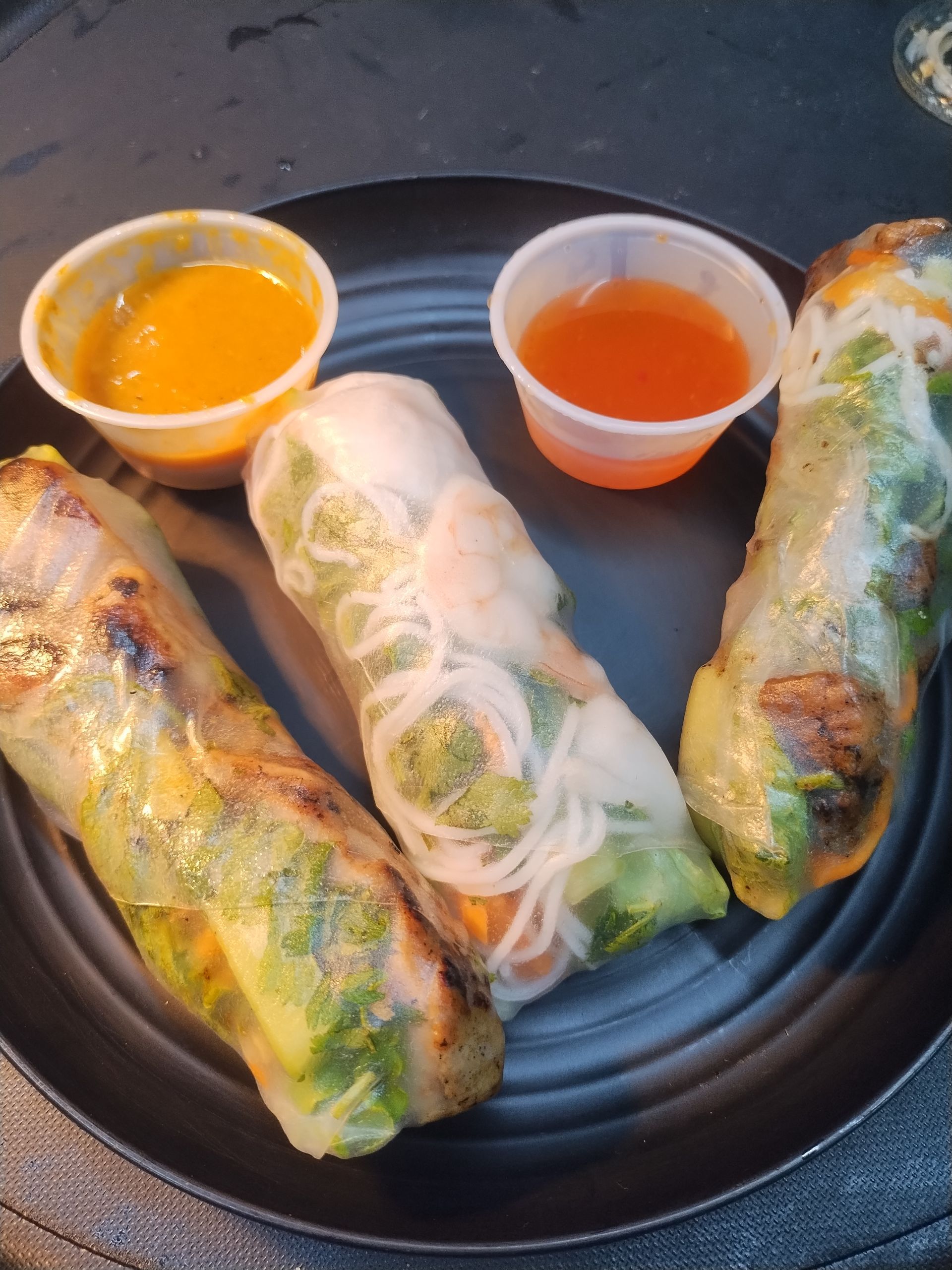 Spring rolls and dipping sauces on a black plate, with one cut open to show noodles and vegetables.
