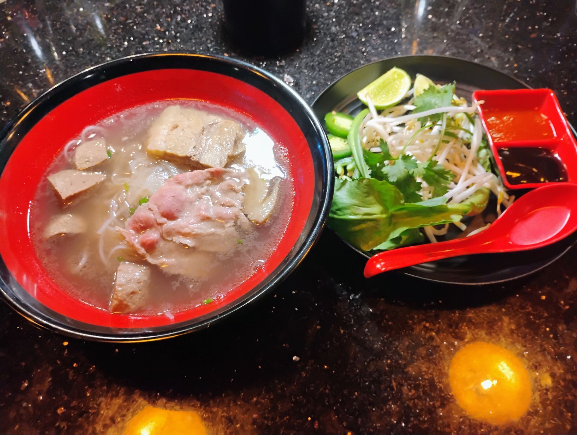Red bowl of noodle soup beside a small salad and dipping sauce on a black table.