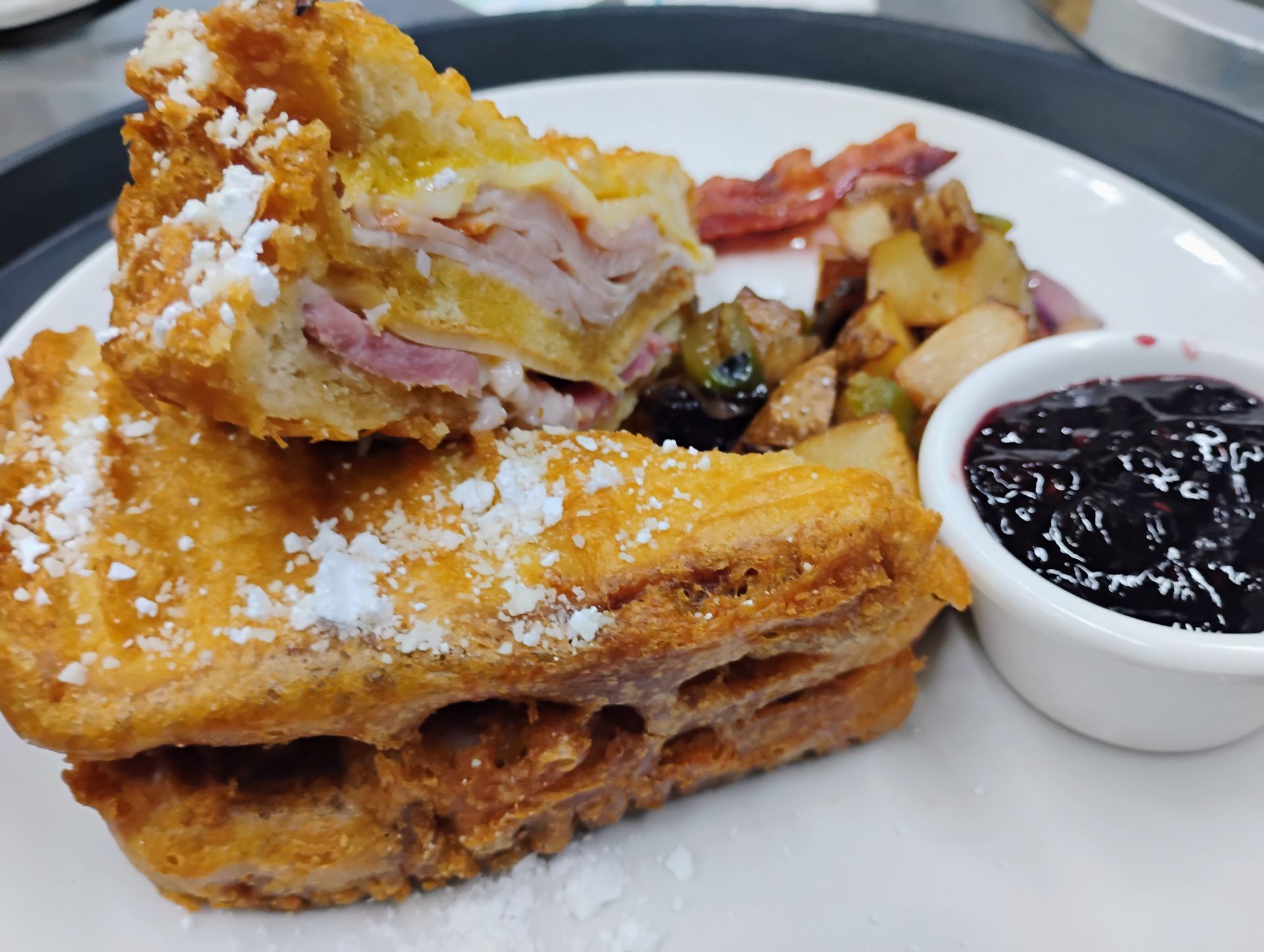 French toast with powdered sugar and jam on a plate, with bacon and fruit on the side