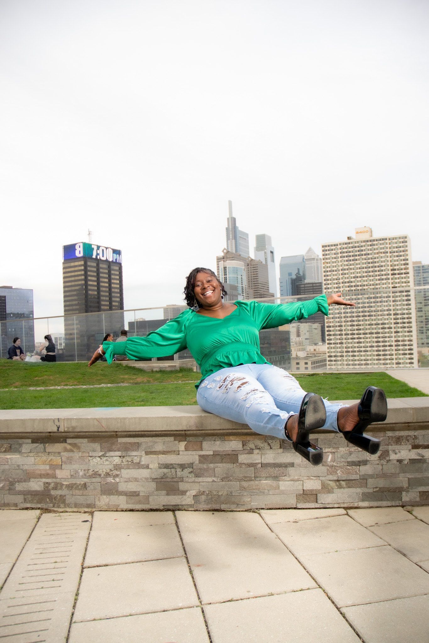 A woman in a green sweater is sitting on a wall with her arms outstretched.