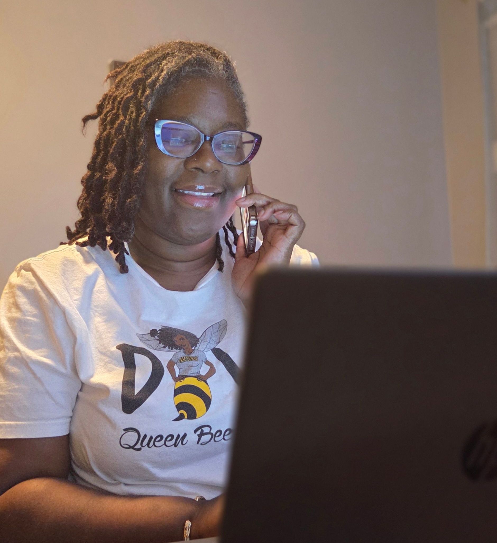 A woman wearing a queen bee shirt is sitting in front of a laptop computer.