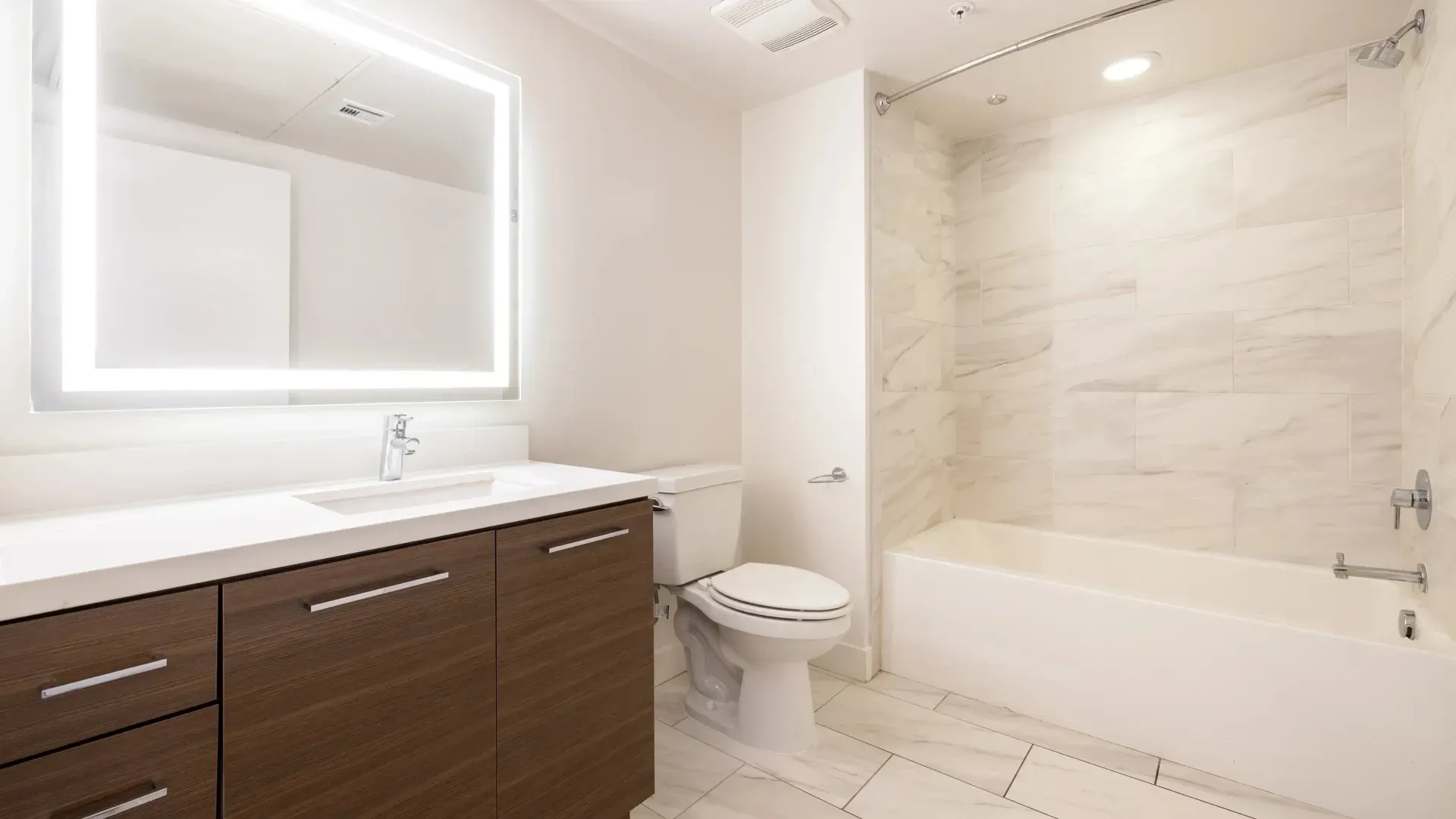 Modern bathroom with a large illuminated mirror, vanity, toilet, and bathtub with marble tile shower surround.