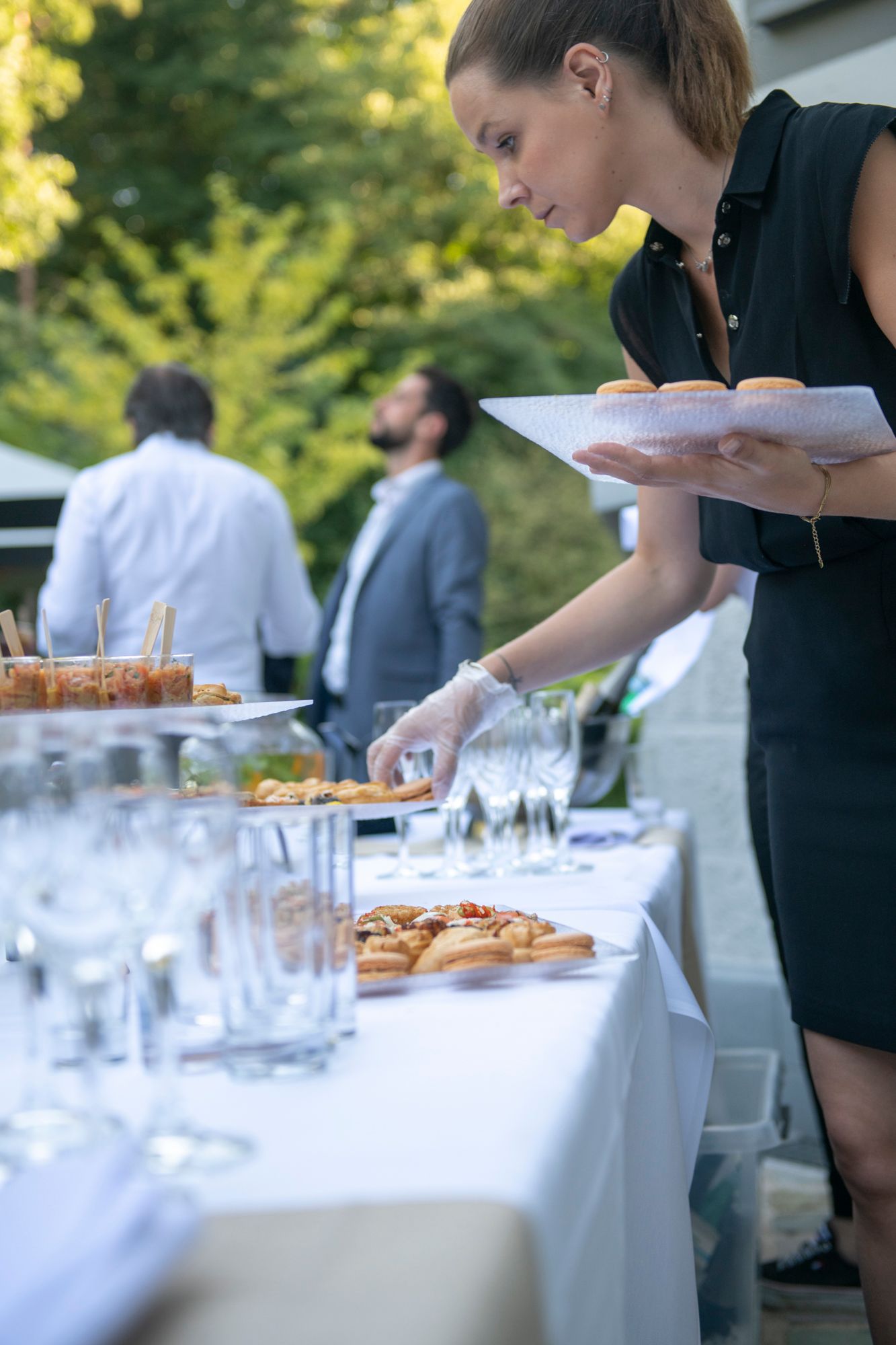 Une femme dispose la nourriture sur un buffet en extérieur. D'autres personnes sont visibles en arrière-plan.