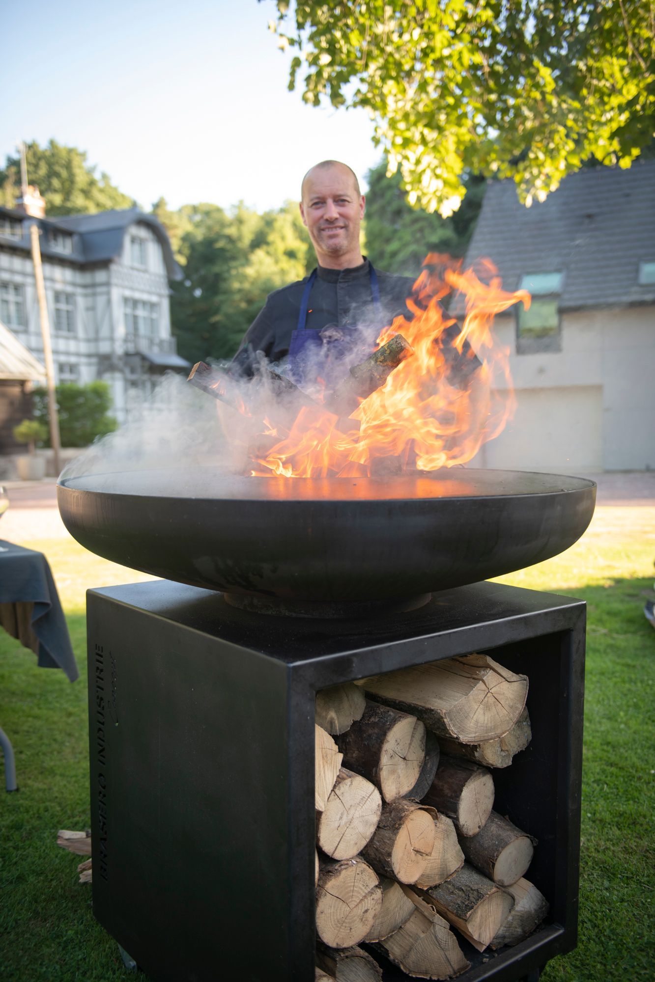 Un homme souriant se tient près d'un foyer extérieur où crépitent des flammes et où s'entremêlent des bûches. Dans un jardin.