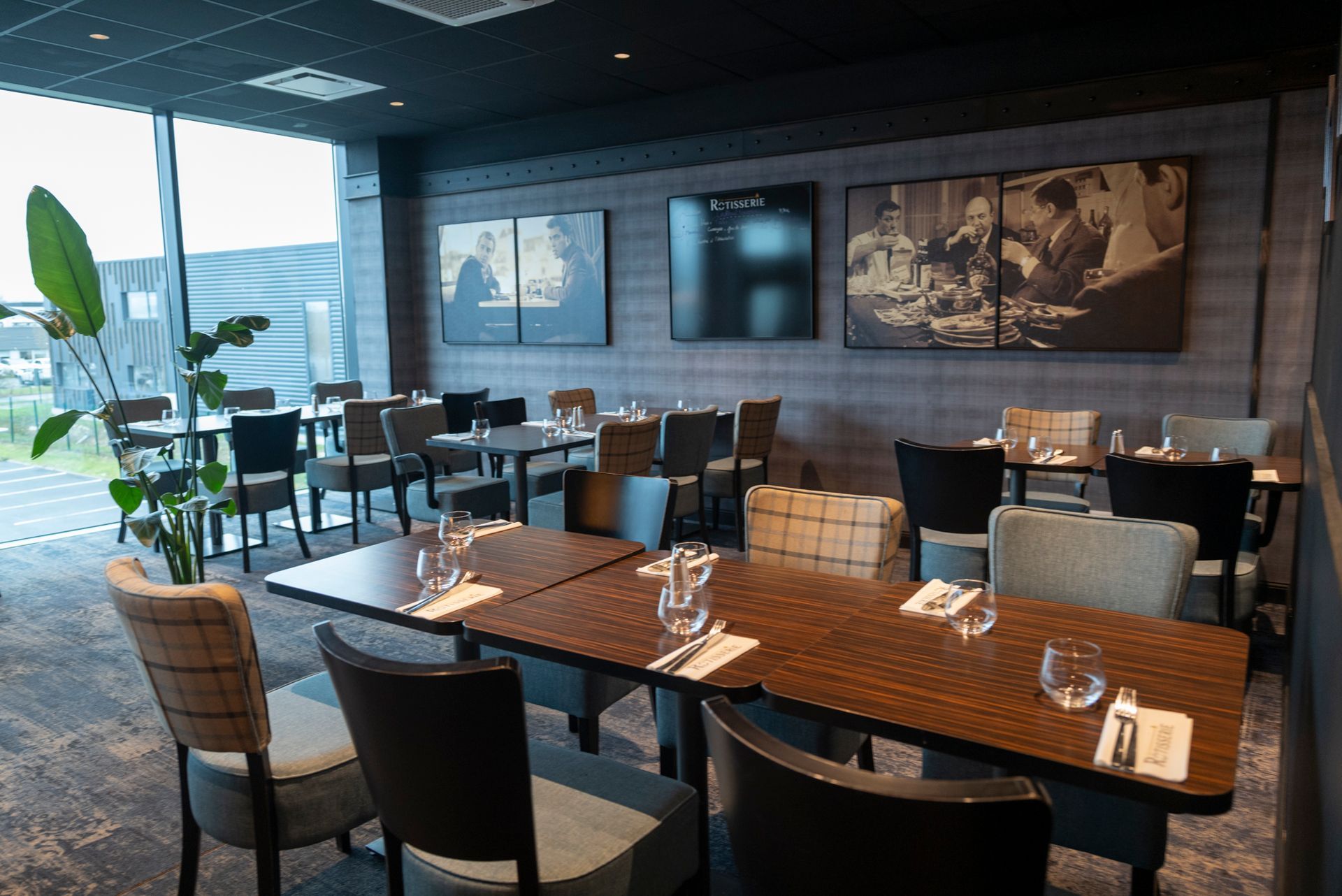 Salle à manger du restaurant avec tables dressées, chaises et œuvres d'art encadrées. Grande fenêtre à gauche.
