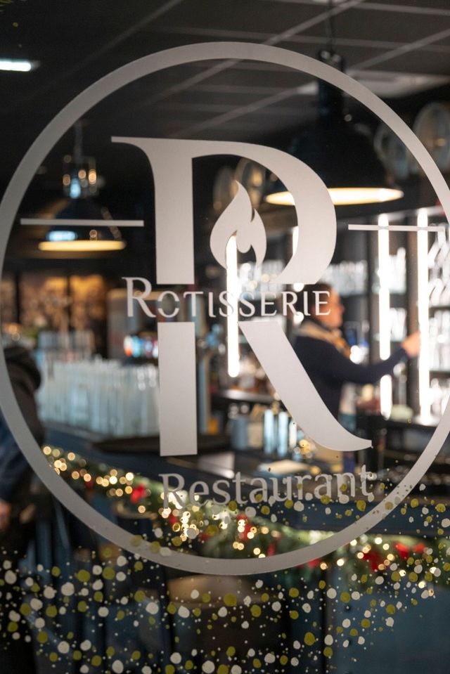 Enseigne du restaurant : grand « R » avec des flammes, « Rôtisserie Restaurant » en dessous, visible à travers une porte vitrée.
