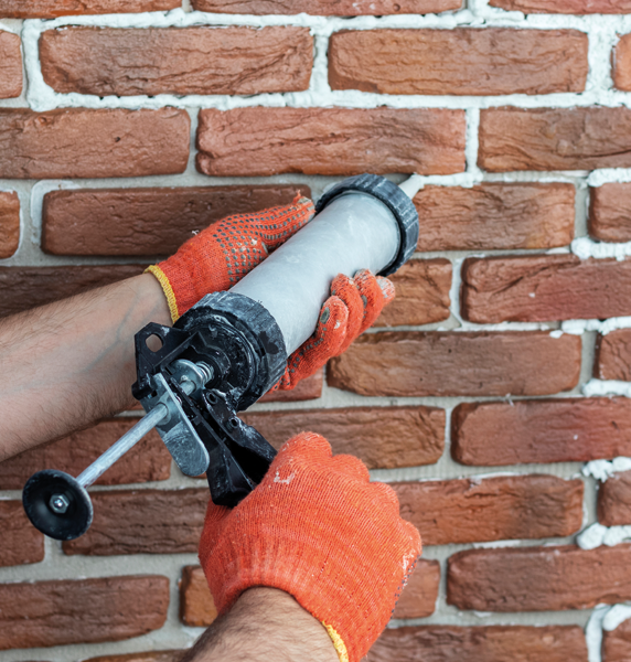A worker in orange gloves uses a caulking gun to apply white mortar between rustic red bricks.